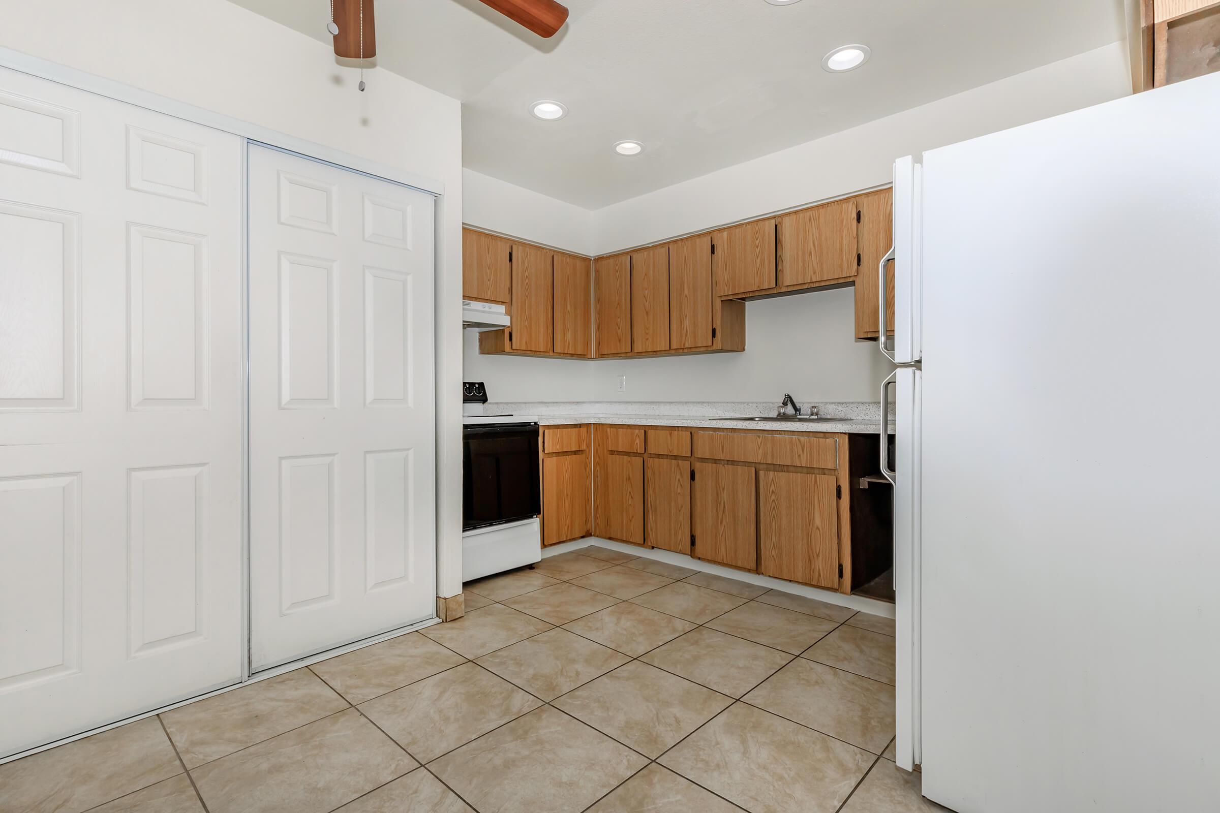 A spacious kitchen featuring wooden cabinets, a white refrigerator, an oven, and a sink. The floor is tiled, and there is a ceiling fan with light fixtures. Additionally, there's a double door pantry on the left side of the image.