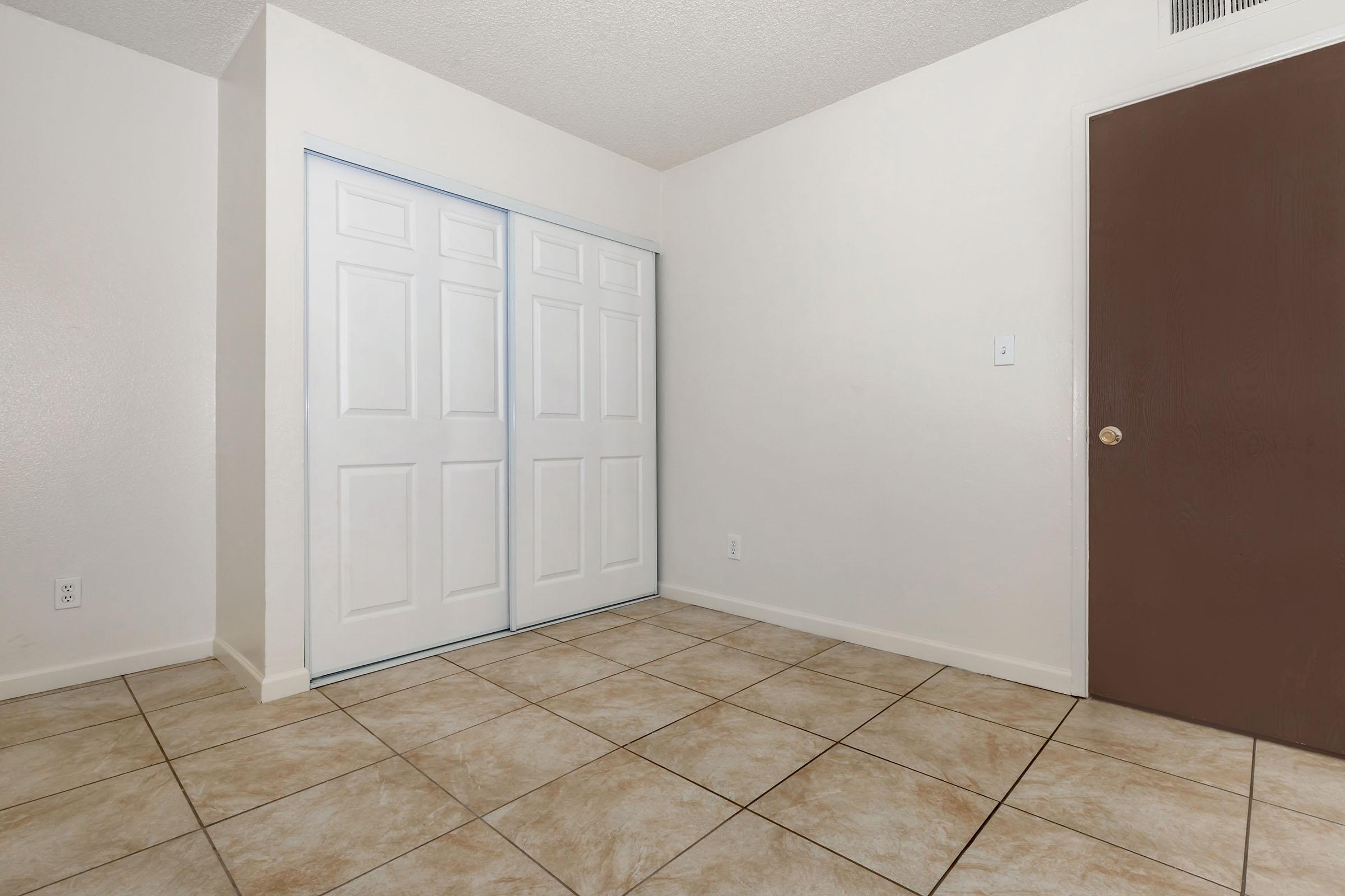 A spacious room featuring a light-colored wall, beige tiled floor, and two closed white sliding doors on the left. A brown door is visible on the right, with no furniture or decorations present, creating an open and empty atmosphere suitable for various uses.