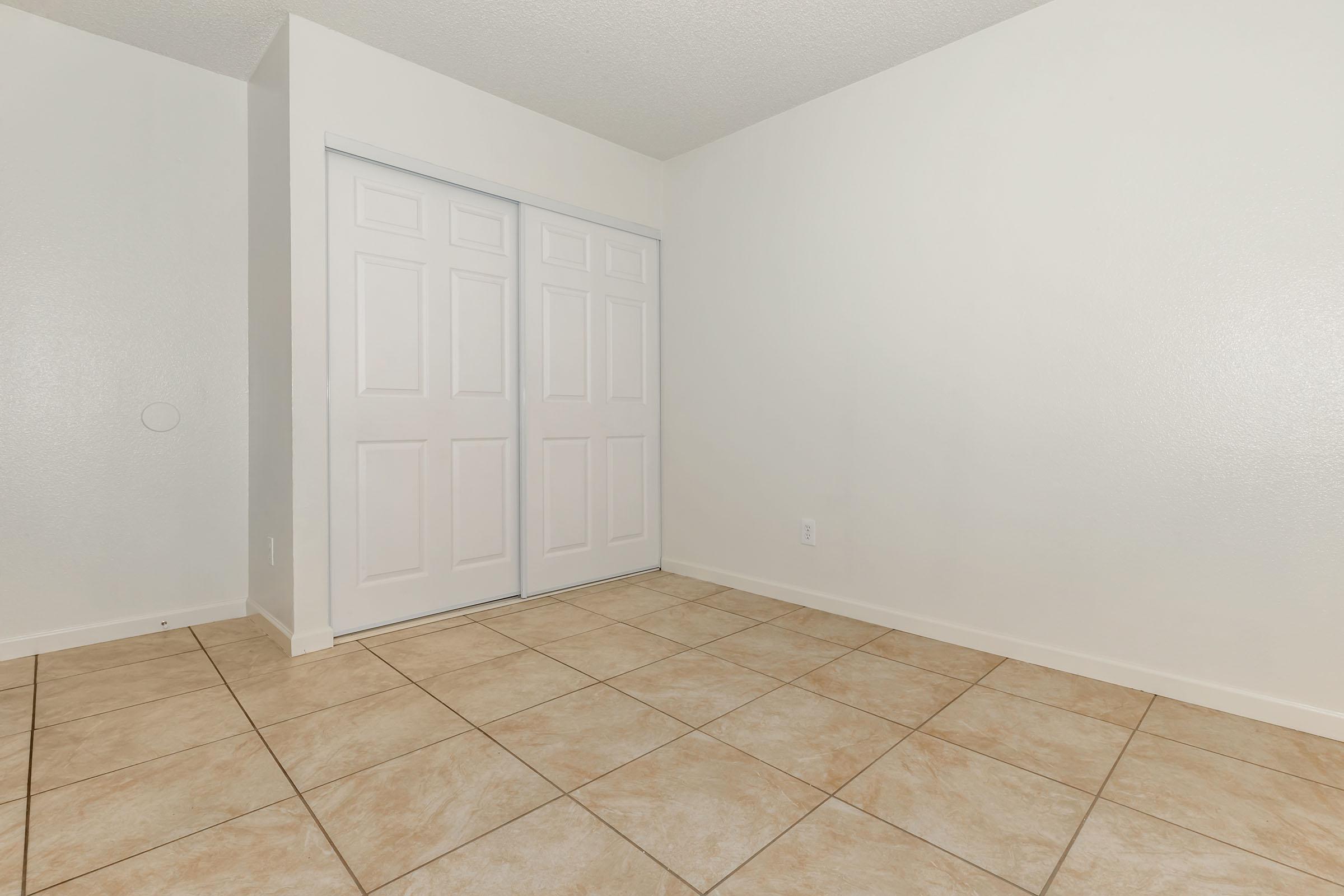 A clean, empty room with white walls and beige tiled flooring. There are sliding closet doors on one wall, and the room appears well-lit and spacious. The overall ambiance is neutral and ready for decoration or furnishings.