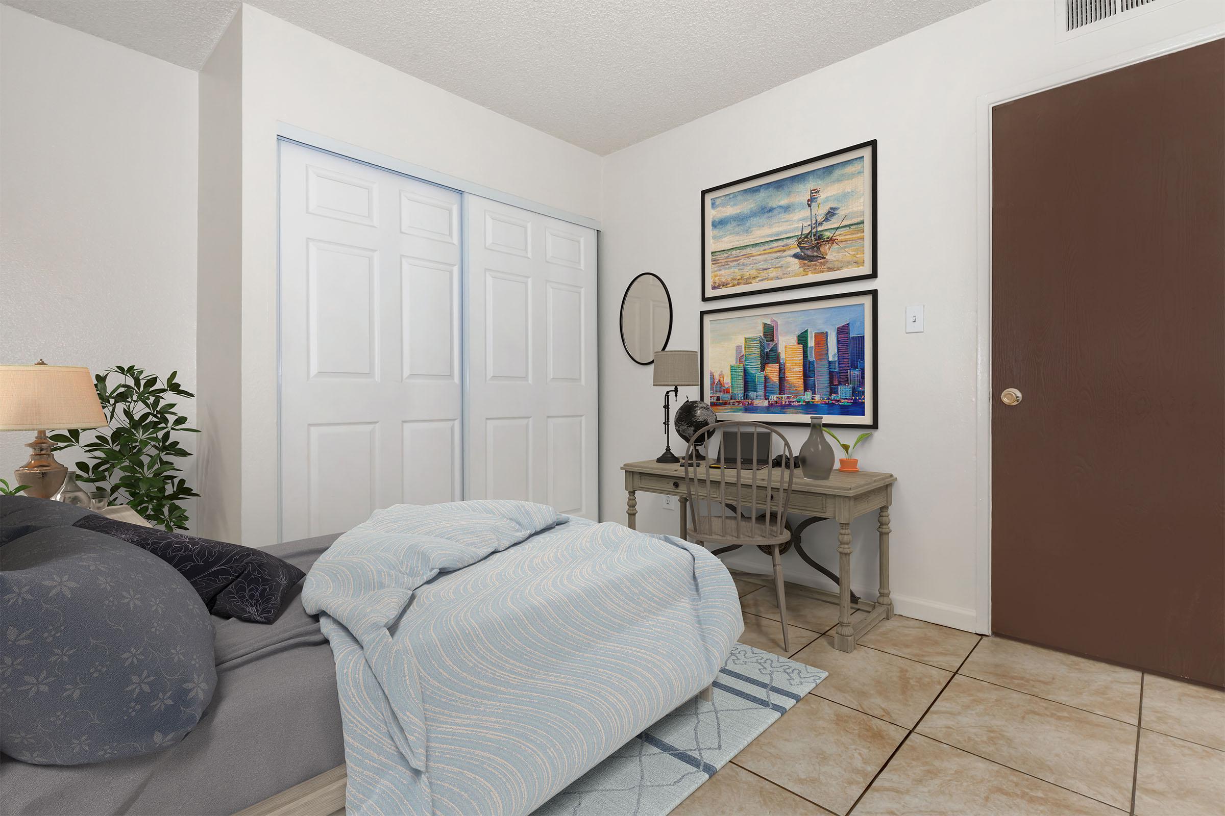 A cozy bedroom featuring a neatly made bed with a blue blanket, a desk and chair with colorful cityscape artwork above, a wall mirror, and a plant. The room has light-colored walls and tiled flooring, with a closed closet door and a brown door leading outside. Soft lighting creates a warm atmosphere.