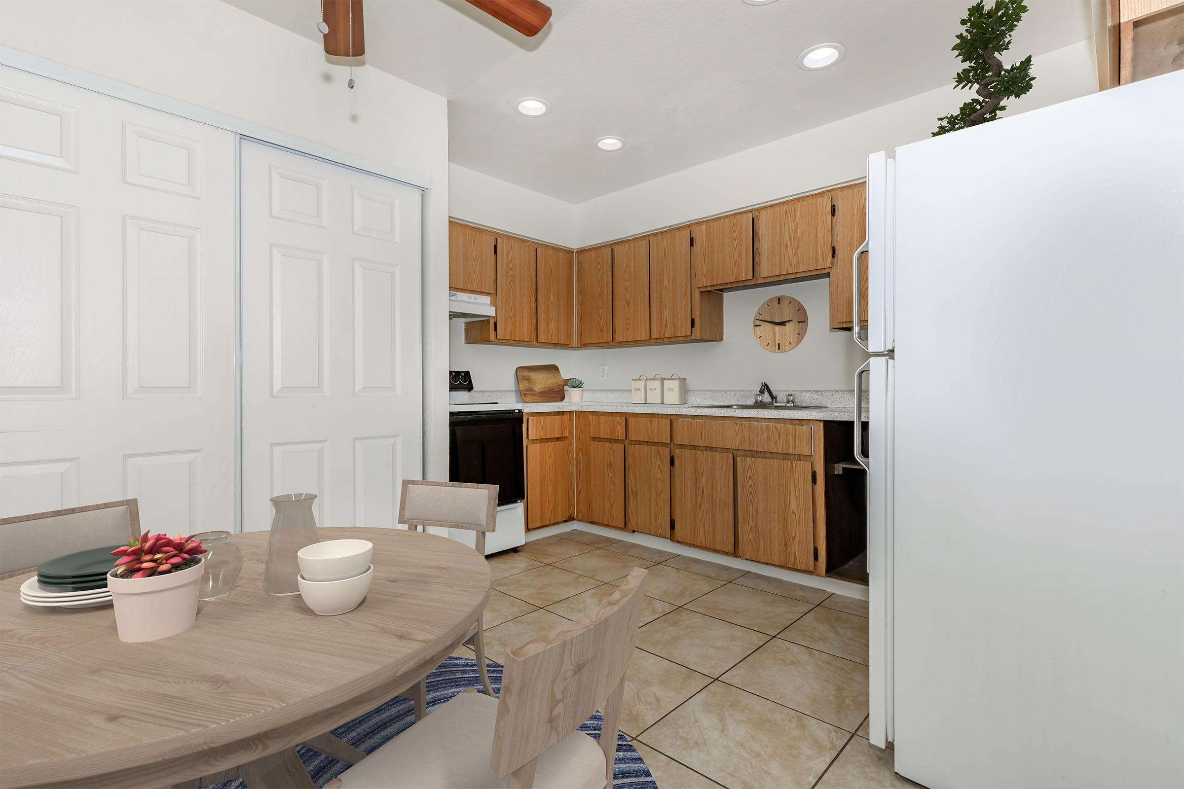 Bright and inviting kitchen featuring wooden cabinets, a spacious countertop, and a stainless steel refrigerator. A round dining table is set with dishes and a small plant. White walls and tiled flooring complement the overall modern aesthetic. Natural light enhances the warm atmosphere.