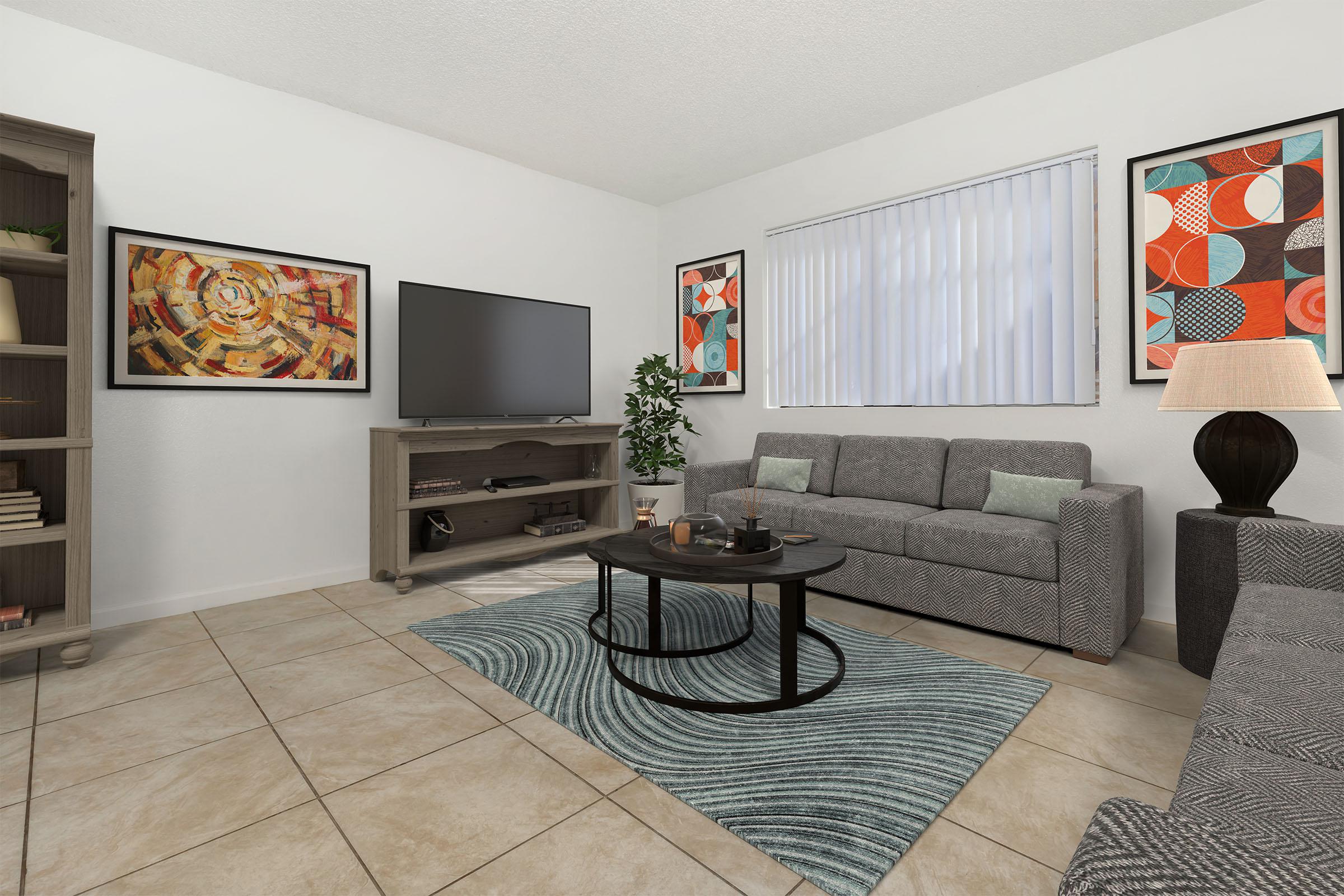 A modern living room featuring a gray sofa and armchair, a round coffee table, and a TV on a console. The walls are decorated with colorful abstract artwork, and there is a potted plant in the corner. The floor is tiled, and a patterned area rug adds texture to the space. Natural light filters through vertical blinds.