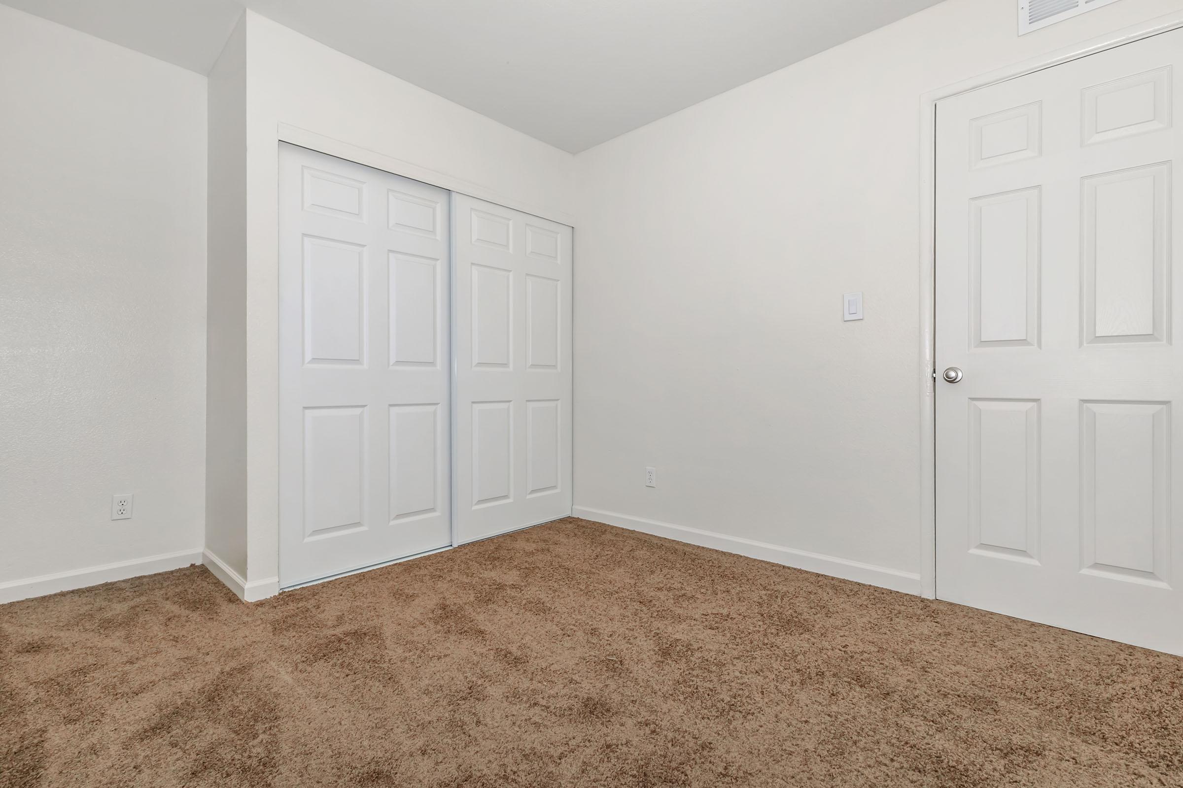 Interior of a vacant room with light-colored walls and a plush, brown carpet. A white double-door closet is visible on one side, and a single white door is on the opposite side. The room is well-lit, creating a spacious and clean ambiance.