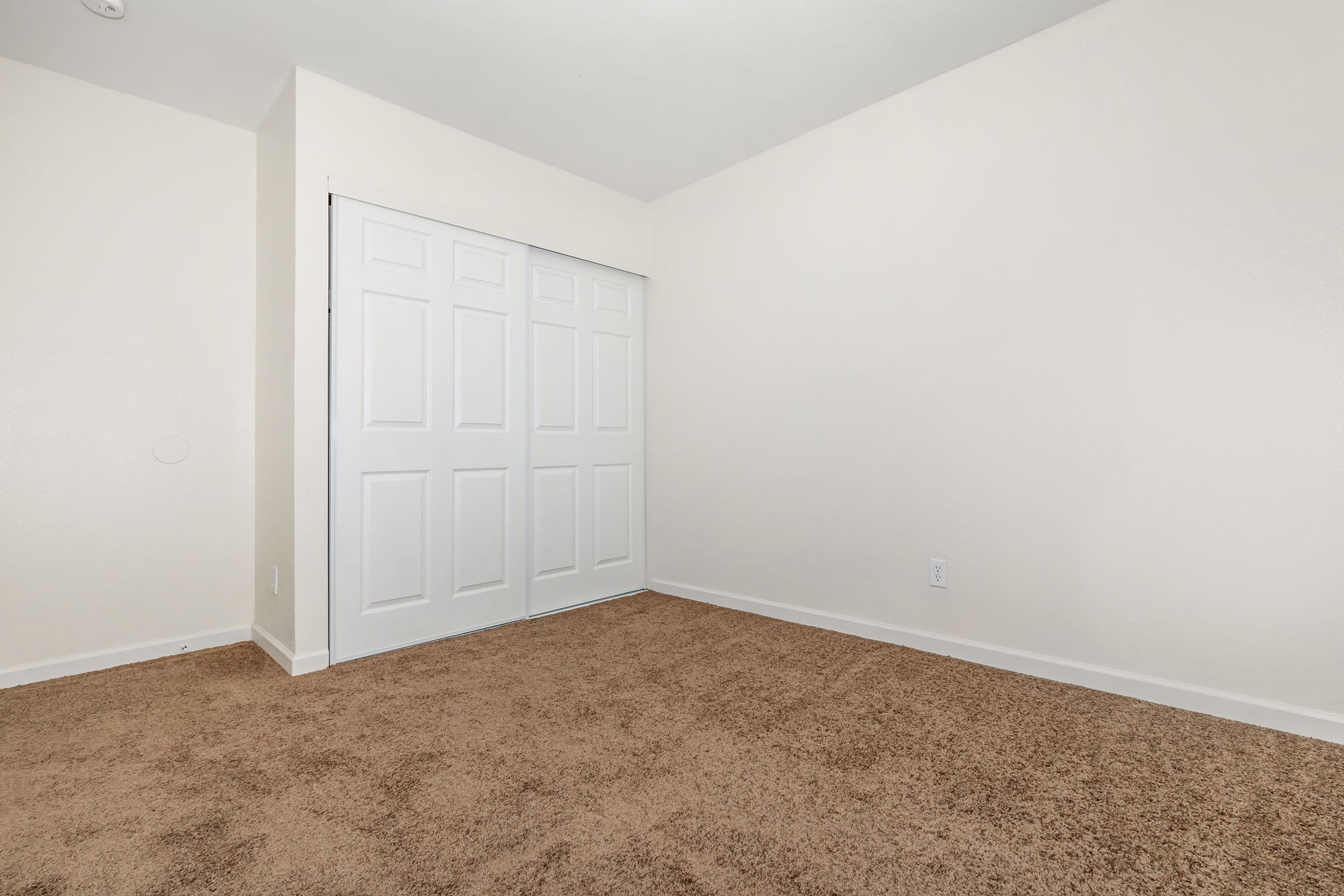 Empty room with light-colored walls and a tan carpet. There is a set of white sliding closet doors on one wall, and the space is well-lit, creating a clean and minimalistic environment.