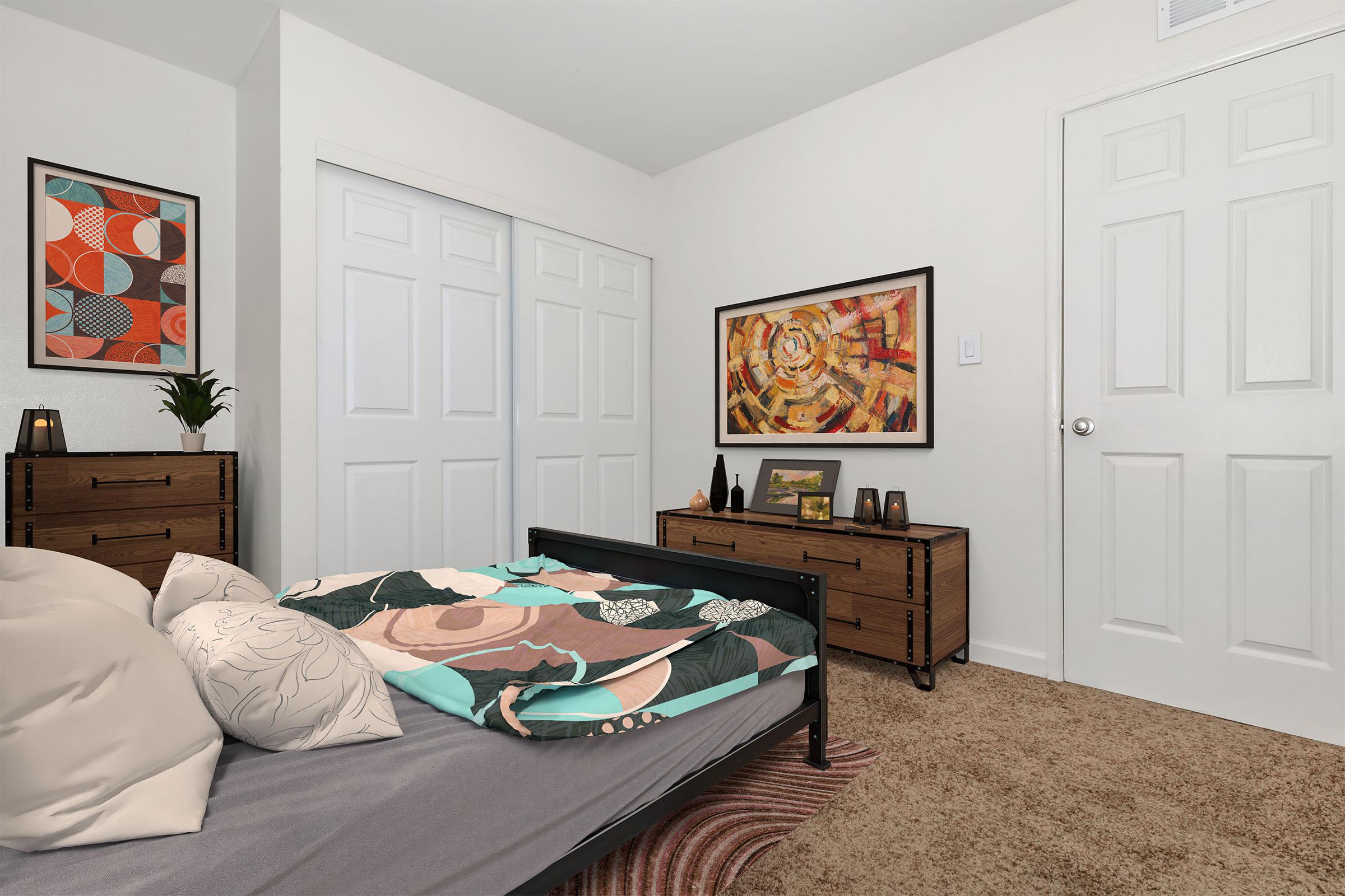 A cozy bedroom featuring a bed with a colorful quilt, modern artwork on the walls, a wooden dresser, and a side table with decorative items. The room has light-colored walls and a plush carpet, creating a warm and inviting atmosphere. Two sliding closet doors can be seen in the background.