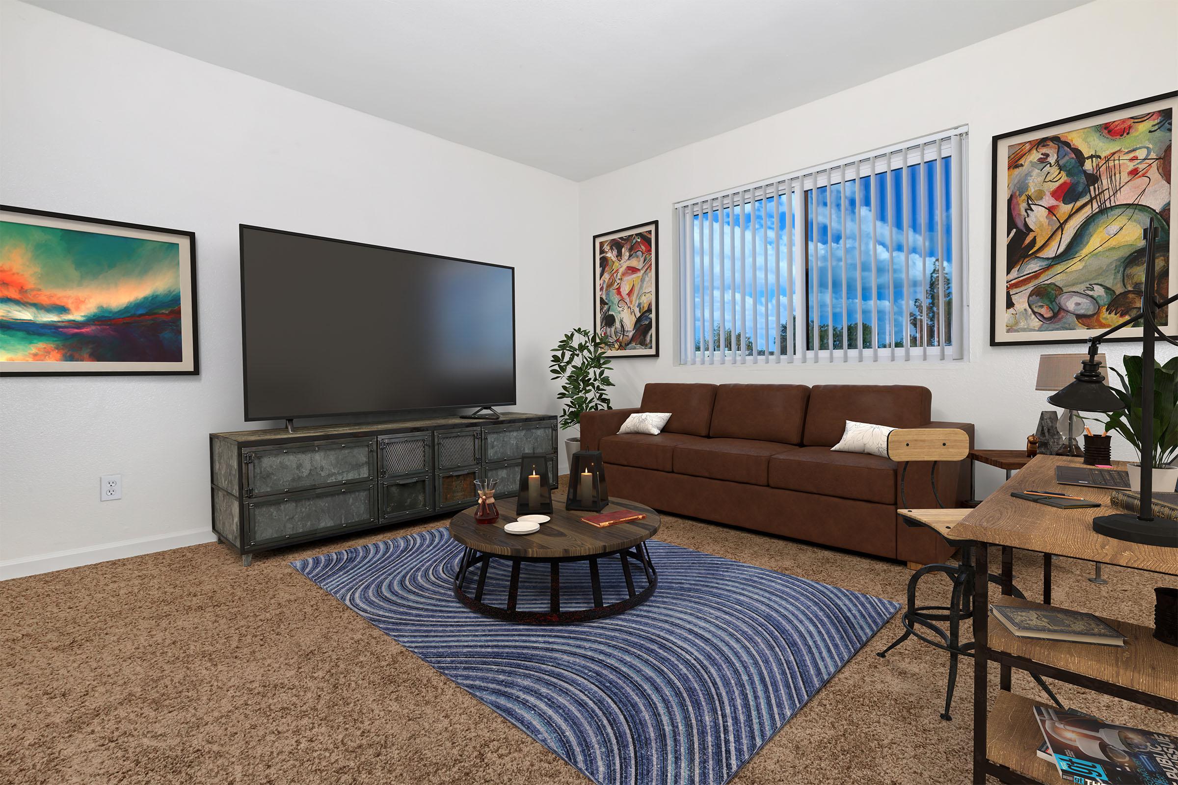 A cozy living room featuring a brown sofa, a large flat-screen TV on a vintage console, and a round coffee table with decorative items. The walls are adorned with colorful artwork, and large windows allow natural light to fill the space. A blue and white patterned rug adds warmth to the carpeted floor.