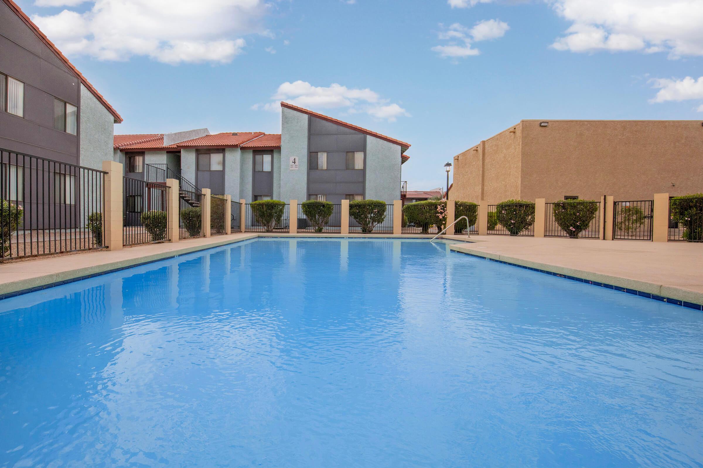 A clear, inviting swimming pool surrounded by a fenced area, with multi-story apartment buildings in the background. The sky is bright blue with a few white clouds, and neatly trimmed bushes line the pool area, creating a serene and welcoming atmosphere.