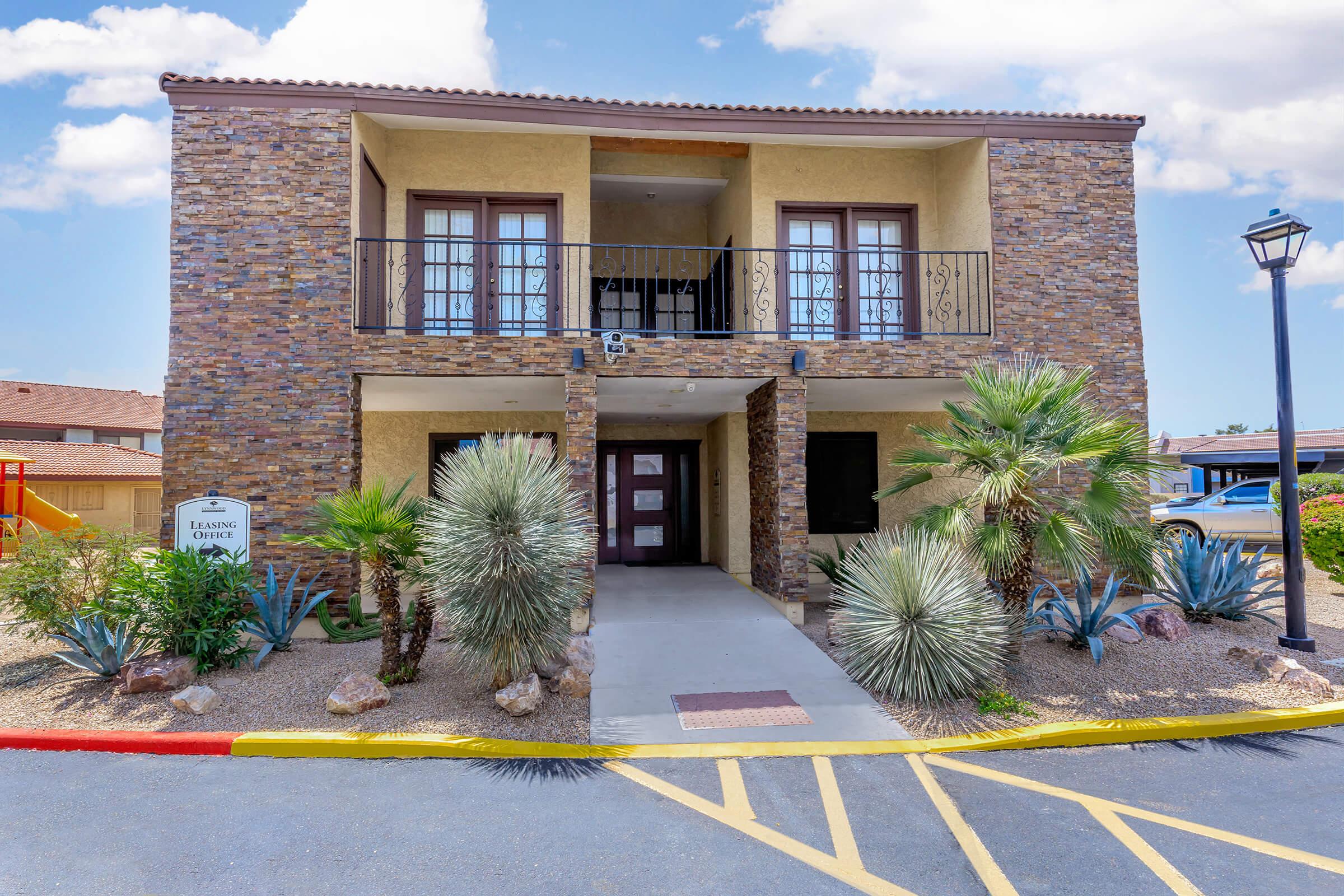 A two-story stone building with a covered entrance, flanked by palm trees and desert plants. The upper balcony features decorative railings, and there are two windows on the second floor. The pathway is lined with colorful paint, leading to the entrance. A streetlamp stands nearby against a bright blue sky.