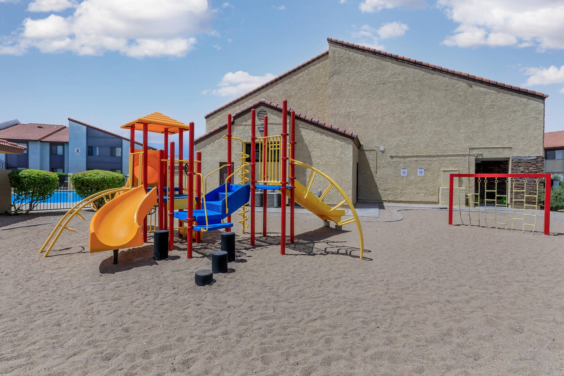 A children's playground featuring colorful slides, climbing structures, and a sandbox area, situated next to a residential building. The sky is clear with a few clouds, and the playground is surrounded by manicured bushes.