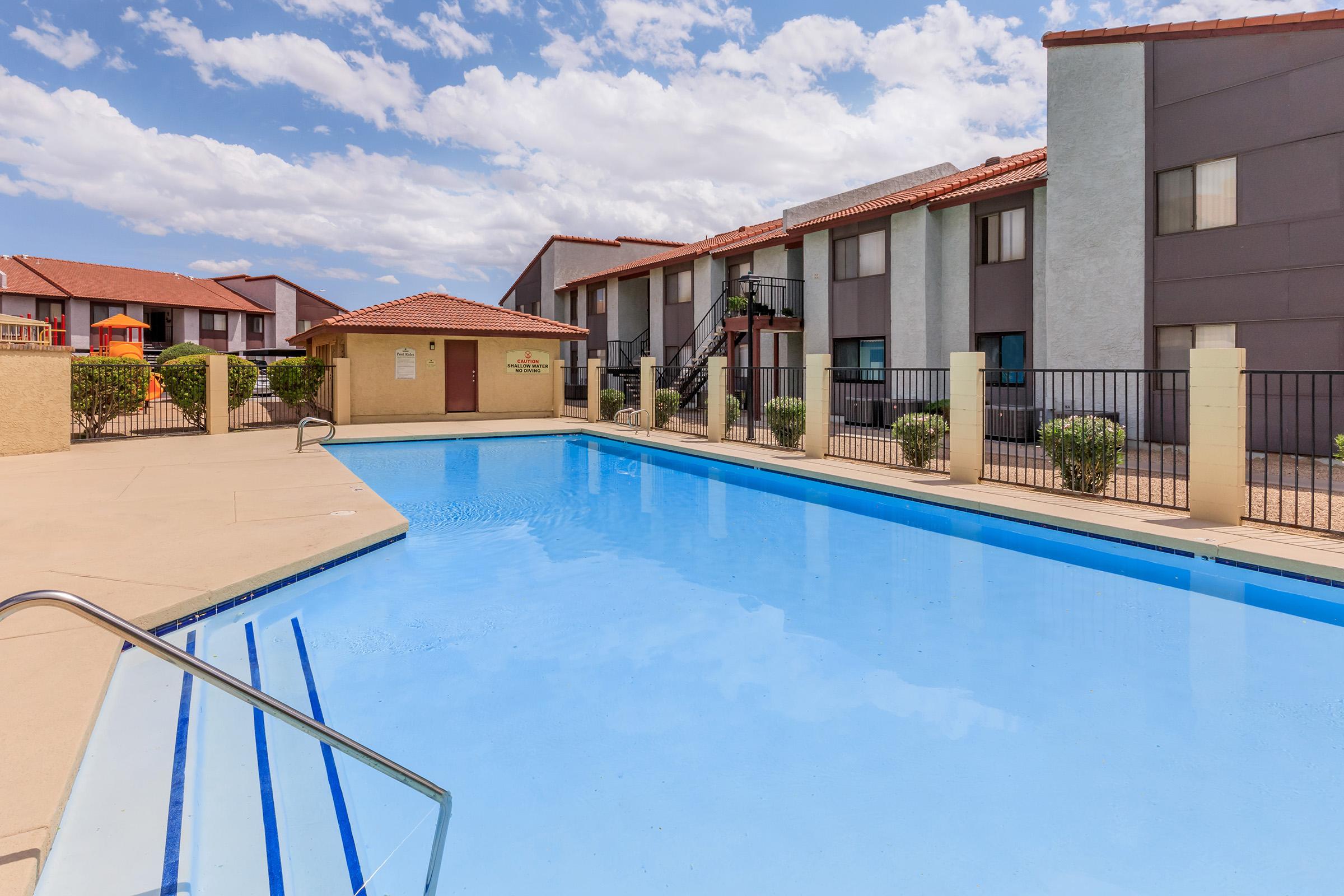 Swimming pool surrounded by a courtyard with modern residential buildings. The pool is clear blue, with a ladder on one side, and there are lounge chairs and patio areas nearby. The sky is partly cloudy, creating a bright and inviting atmosphere.