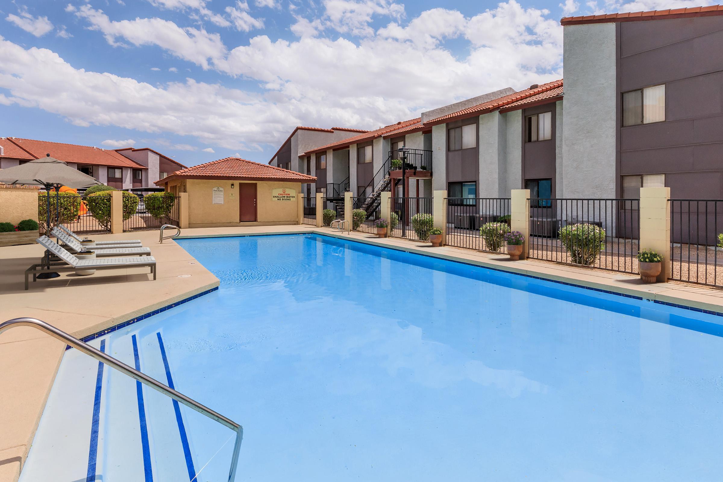 A clear blue swimming pool surrounded by lounge chairs, with a backdrop of two-story apartment buildings. The sky is partly cloudy, and there are potted plants along the pool deck. A shaded seating area is visible in the background, contributing to a relaxed atmosphere.