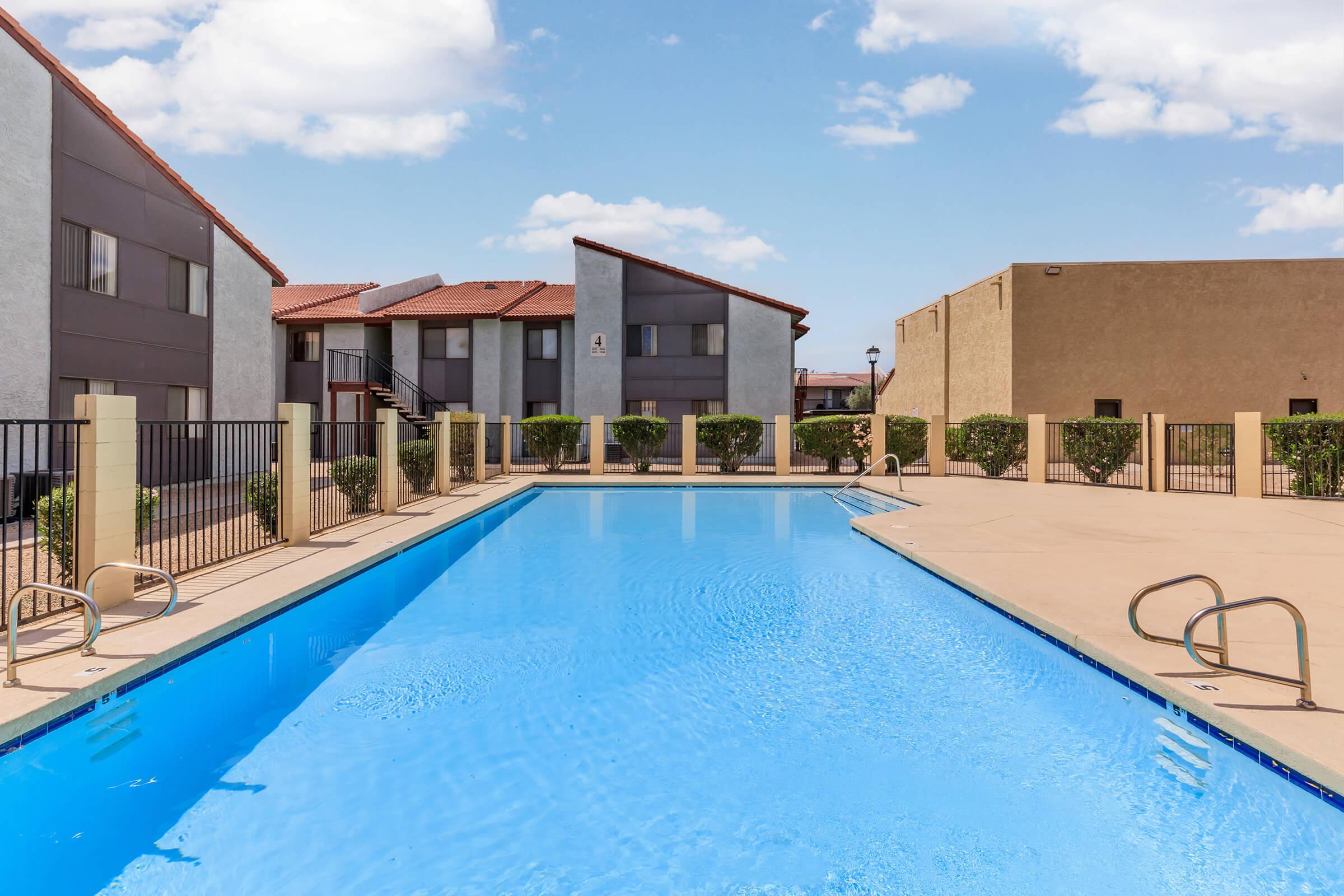 Swimming pool surrounded by a fenced area, with several apartment buildings in the background. The pool water is clear and blue, and there are neatly trimmed shrubs along the pool's edge. The sky is partly cloudy, adding to the inviting atmosphere of the scene.