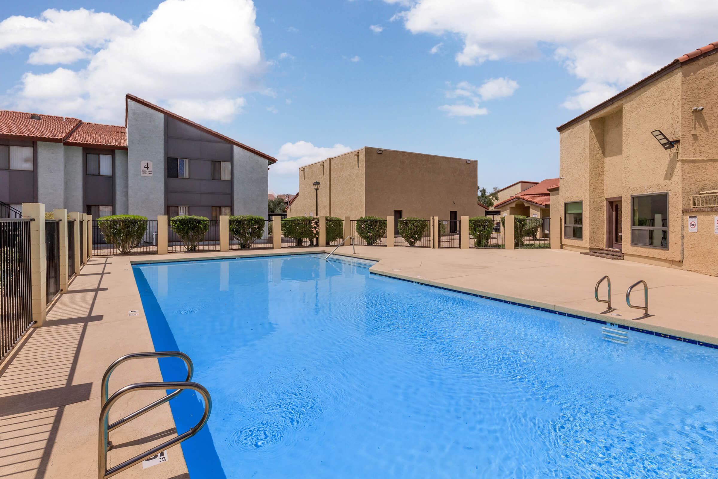 A clear blue swimming pool surrounded by a paved area and a fence. In the background, there are beige apartment buildings with tiled roofs and neatly trimmed shrubs. The sky is bright with a few white clouds.
