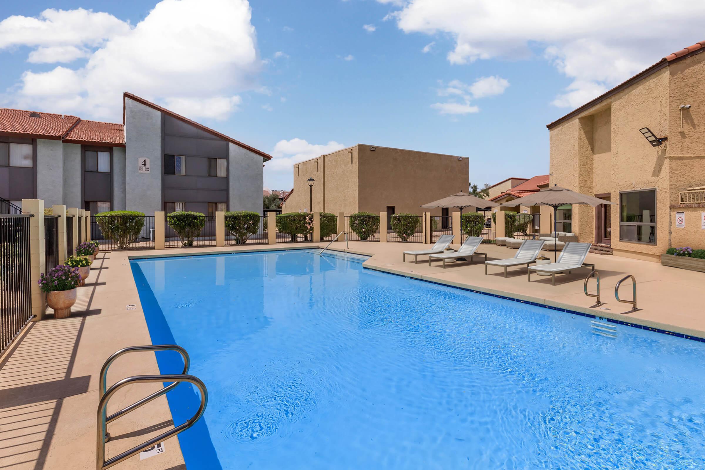 A clear blue swimming pool surrounded by lounge chairs, with a fenced area. There are two buildings in the background, one with a beige exterior and the other with grey, along with lush bushes lining the pool area. The sky is bright and partly cloudy.