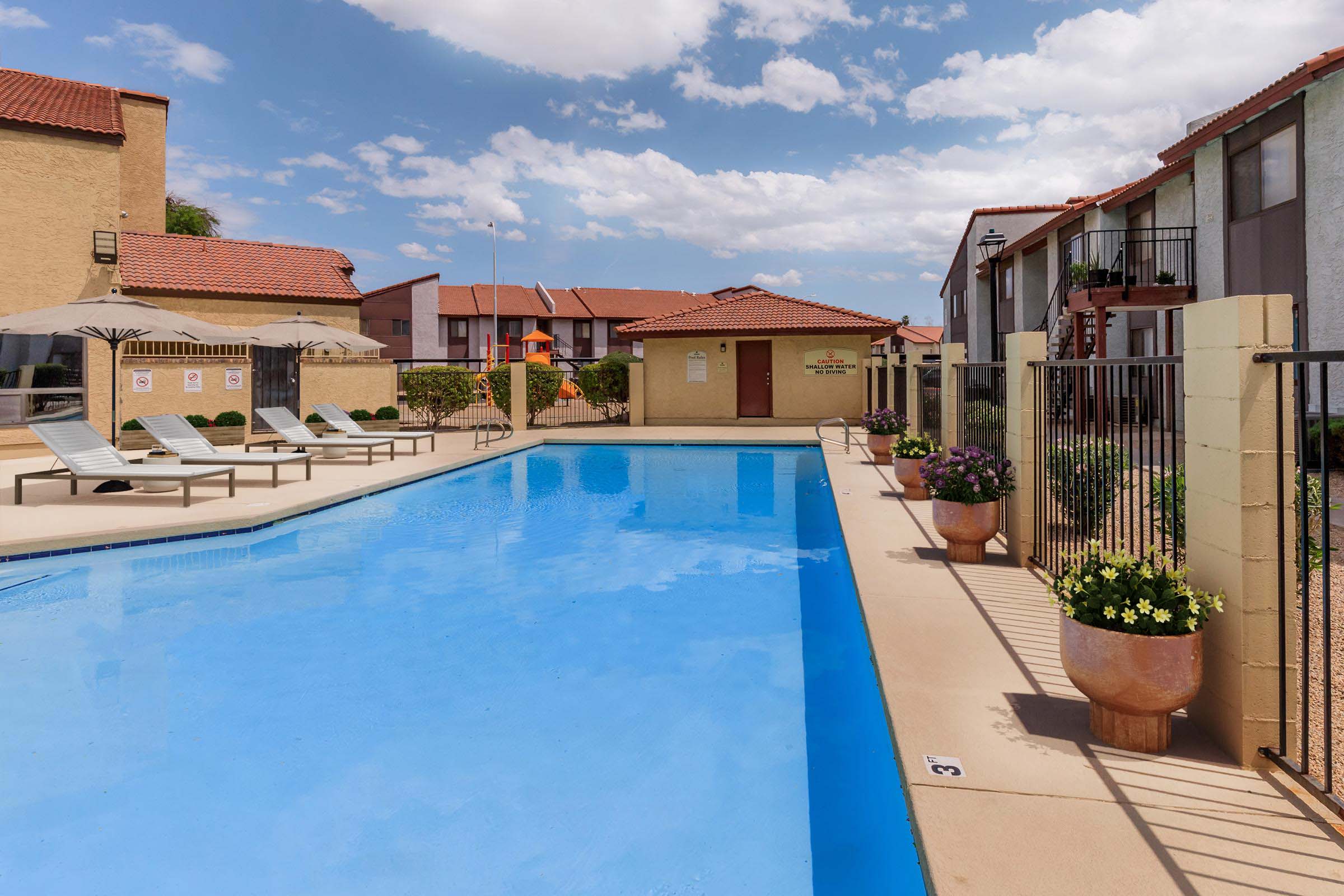 A bright pool area with clear blue water surrounded by lounge chairs and potted plants. In the background, there are several two-story buildings with red roofs under a partly cloudy sky. The area appears clean and inviting, perfect for relaxation or leisure activities.