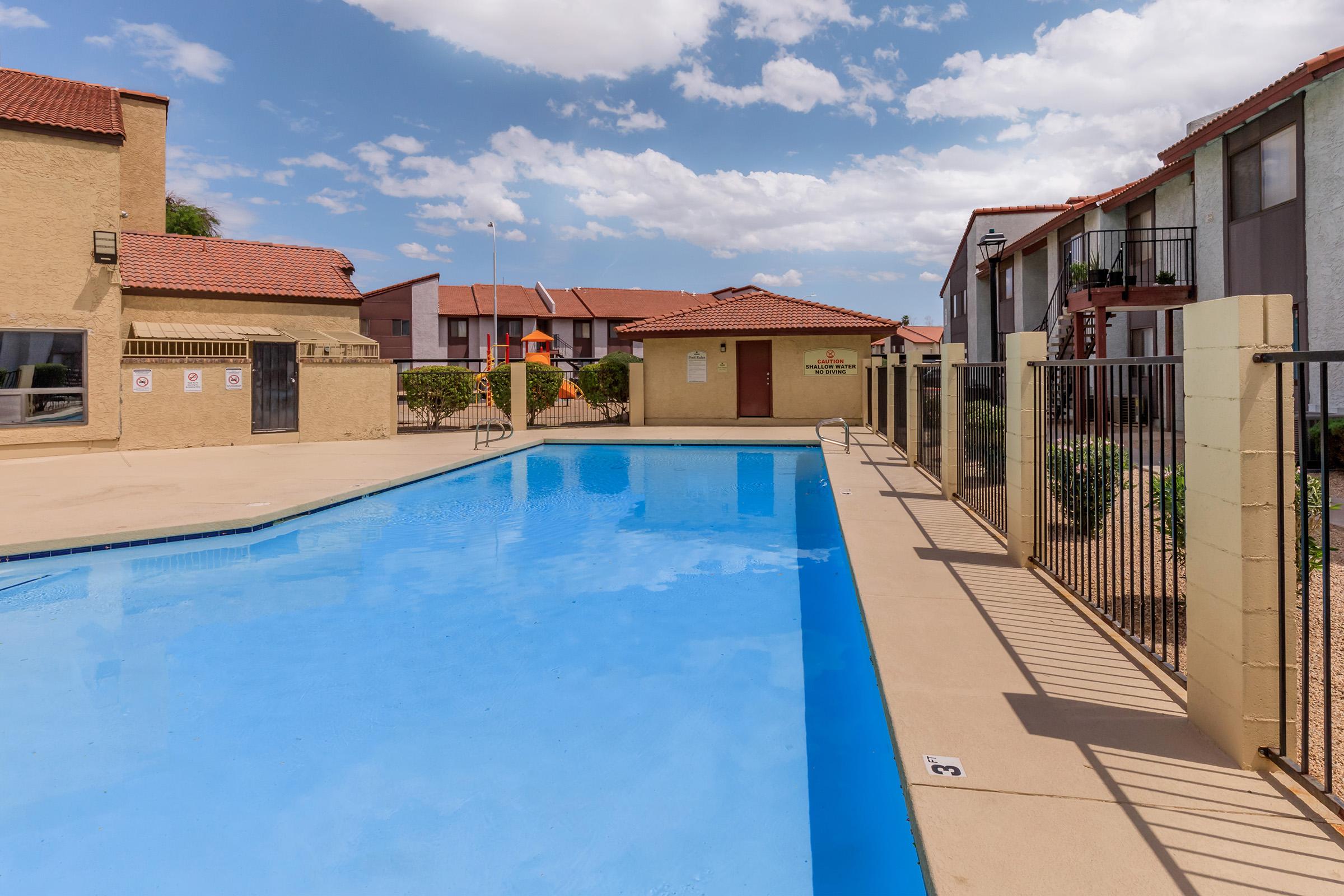 A clear blue swimming pool surrounded by beige buildings with red-tiled roofs. The pool area features a fenced perimeter and lounge space, with a few potted plants. In the background, there are multiple residential units and a partly cloudy sky.