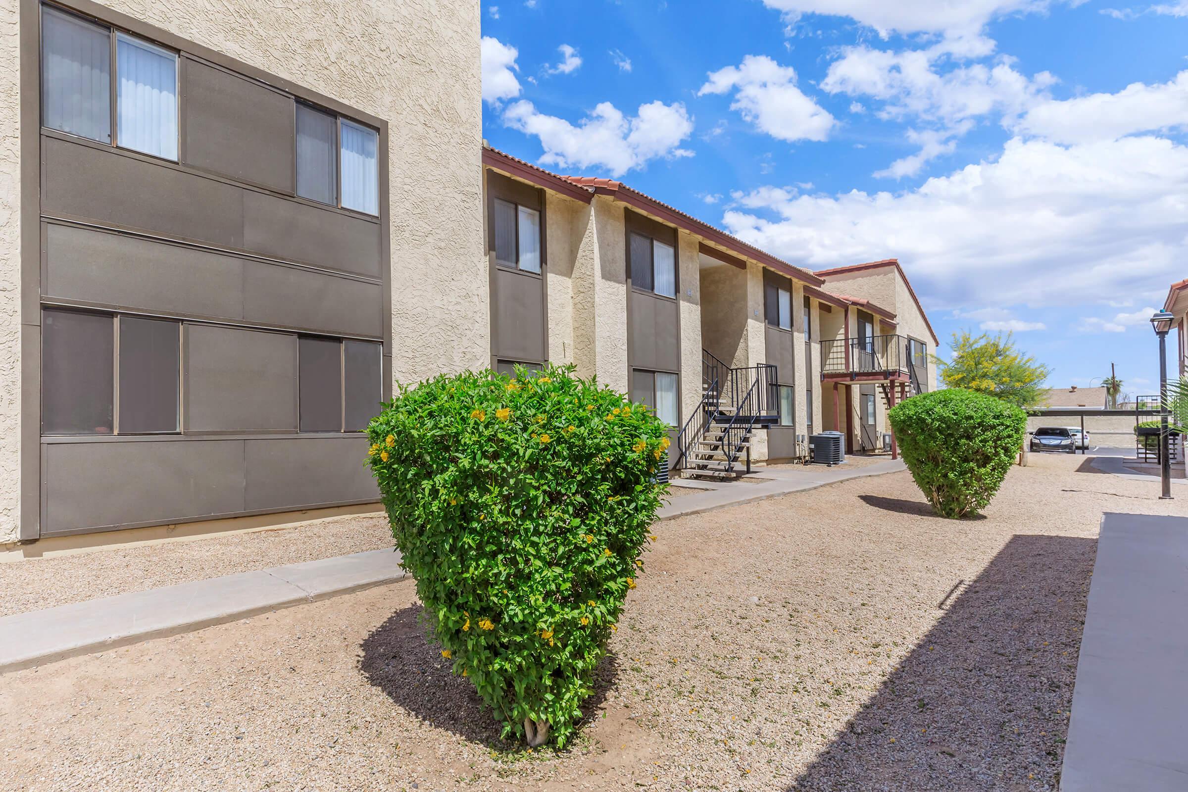 Exterior view of an apartment complex featuring two-story buildings with gray siding, surrounded by well-maintained shrubs and gravel pathways. The sky is partly cloudy, and there are stairs leading to the upper floor entrances. Sunlight illuminates the scene, creating a bright atmosphere.