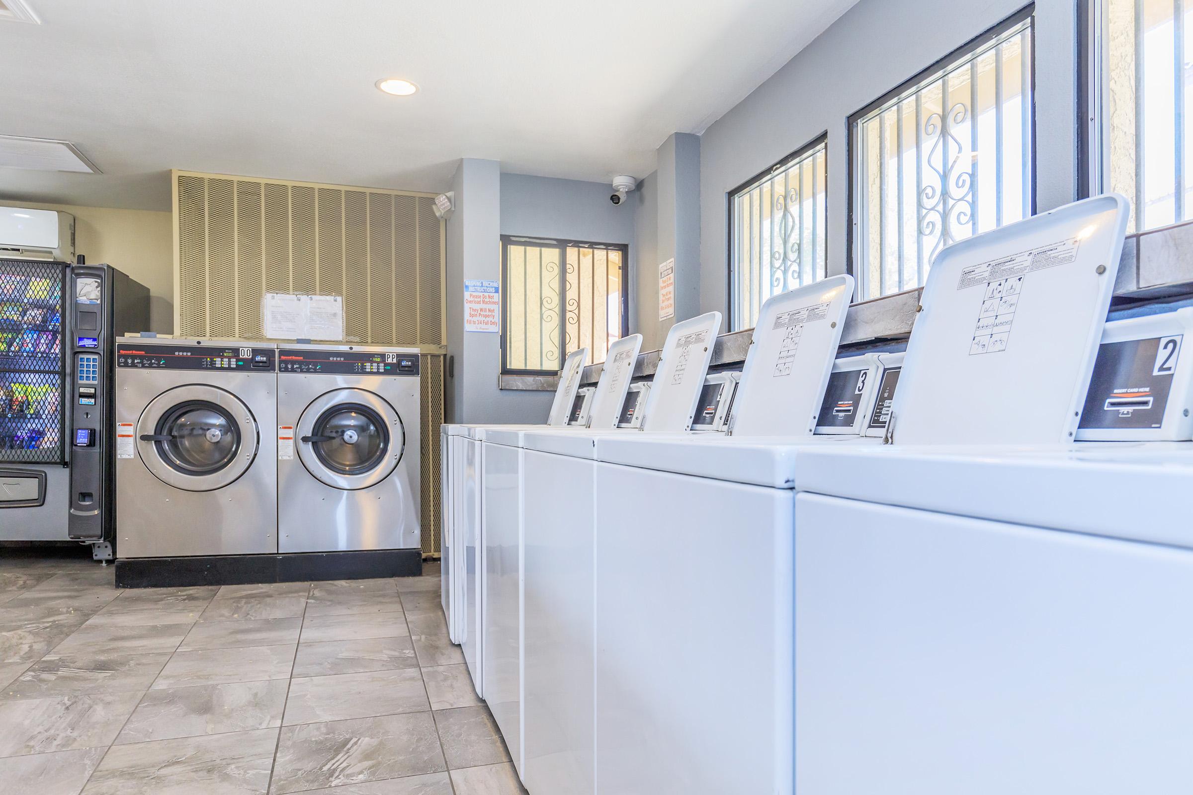 A clean and bright laundry facility featuring several washing machines and dryers lined up against the wall. There is a vending machine in the corner, and large windows allow natural light to flood the space. The flooring is modern and tiled, creating a welcoming atmosphere for patrons.