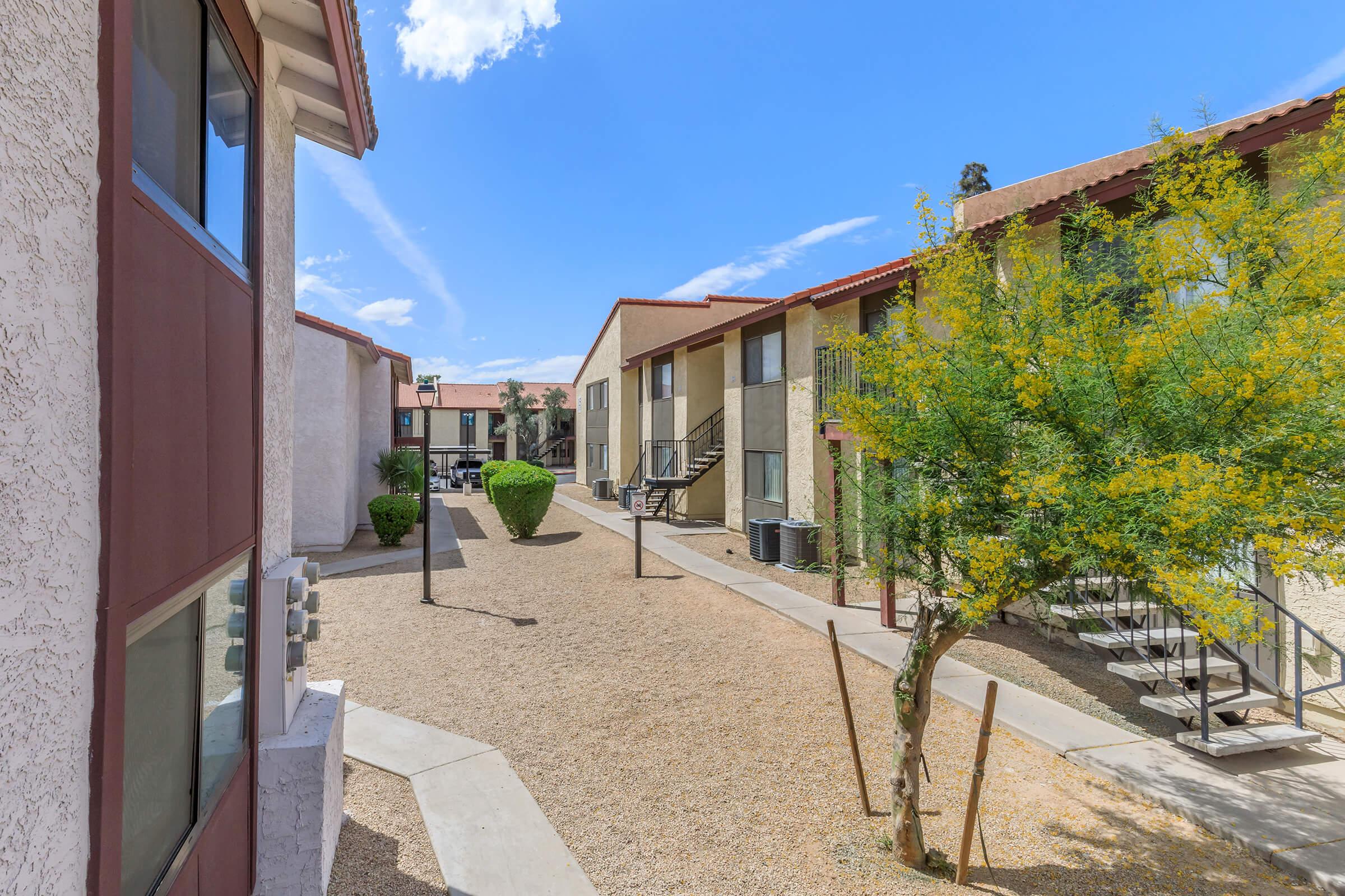 A view of an apartment complex courtyard featuring two-story buildings with beige and brown exteriors. The area has gravel landscaping, small bushes, and a tree, under a clear blue sky with a few clouds. The scene conveys a quiet residential environment.
