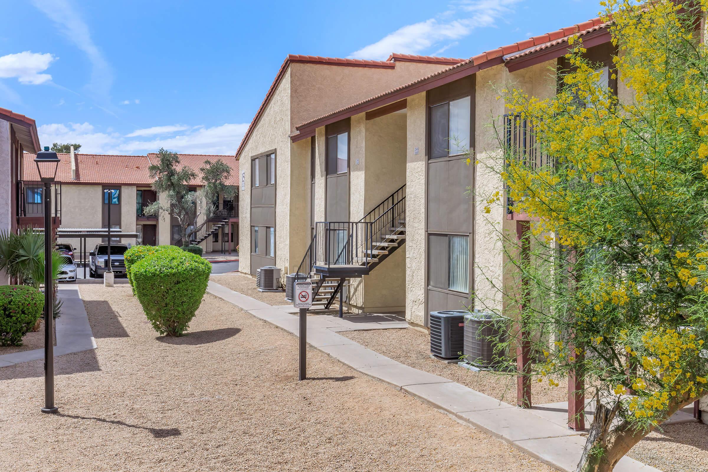 View of a residential apartment complex featuring two-story buildings with balconies, surrounded by landscaped areas with shrubs and trees. A pathway leads through the center, and utility units are visible outside the buildings under a clear blue sky.