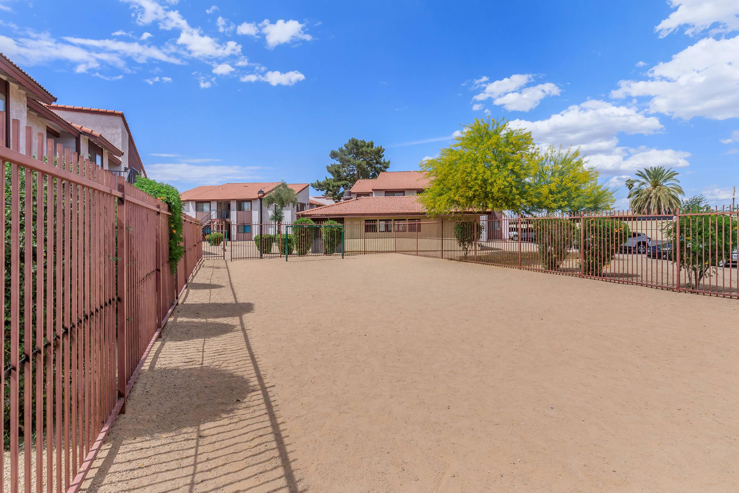 A spacious outdoor area enclosed by a fence, featuring sandy ground and a few small trees. Background includes two-story buildings with red roofs under a clear blue sky with scattered clouds. Sunlight casts shadows along the fence.