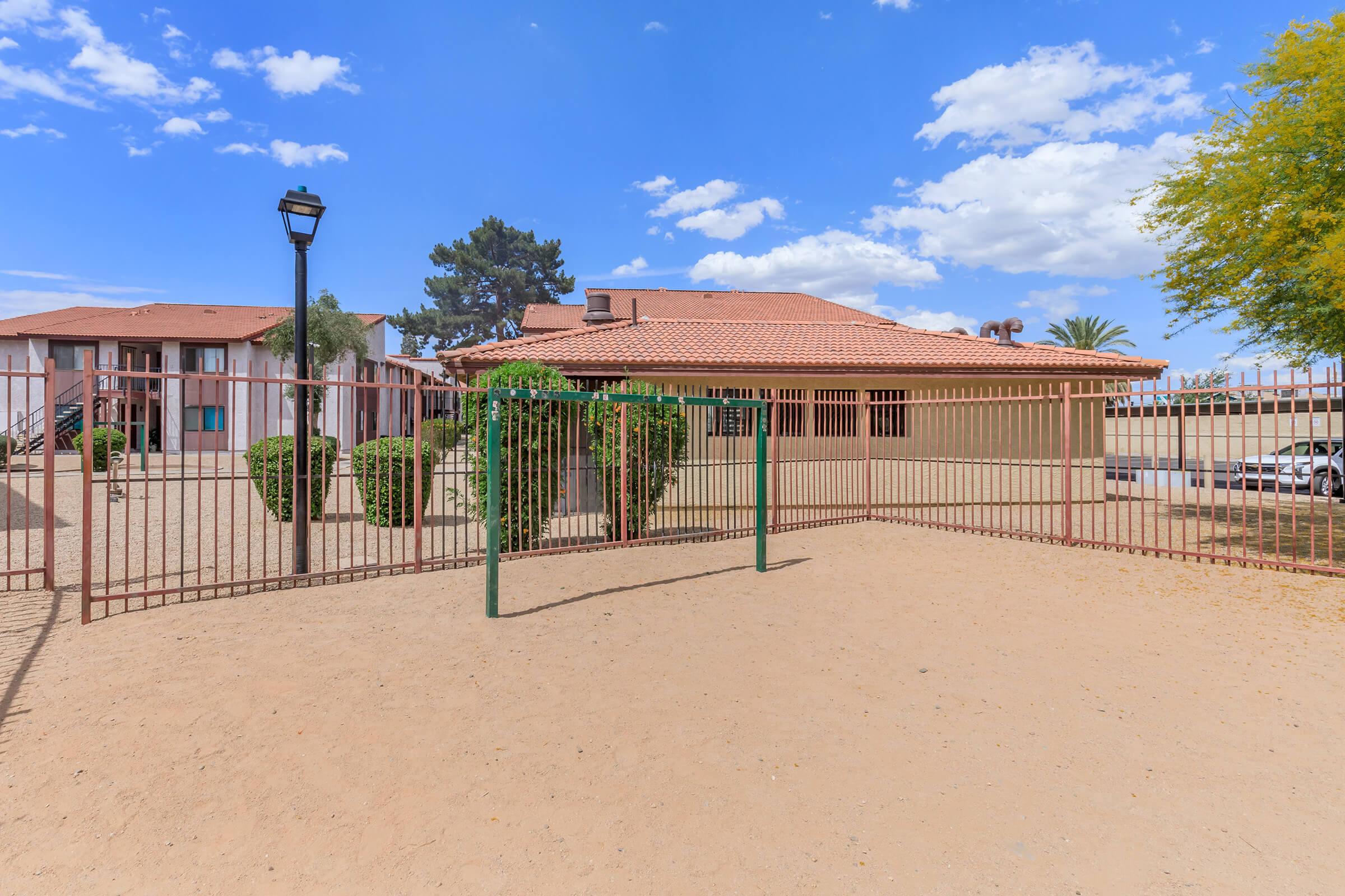 A fenced area surrounding a building with a reddish-brown roof, featuring a gravel driveway. Green shrubs are visible within the fenced space, and several trees are in the background. The sky is partly cloudy with blue and white clouds.