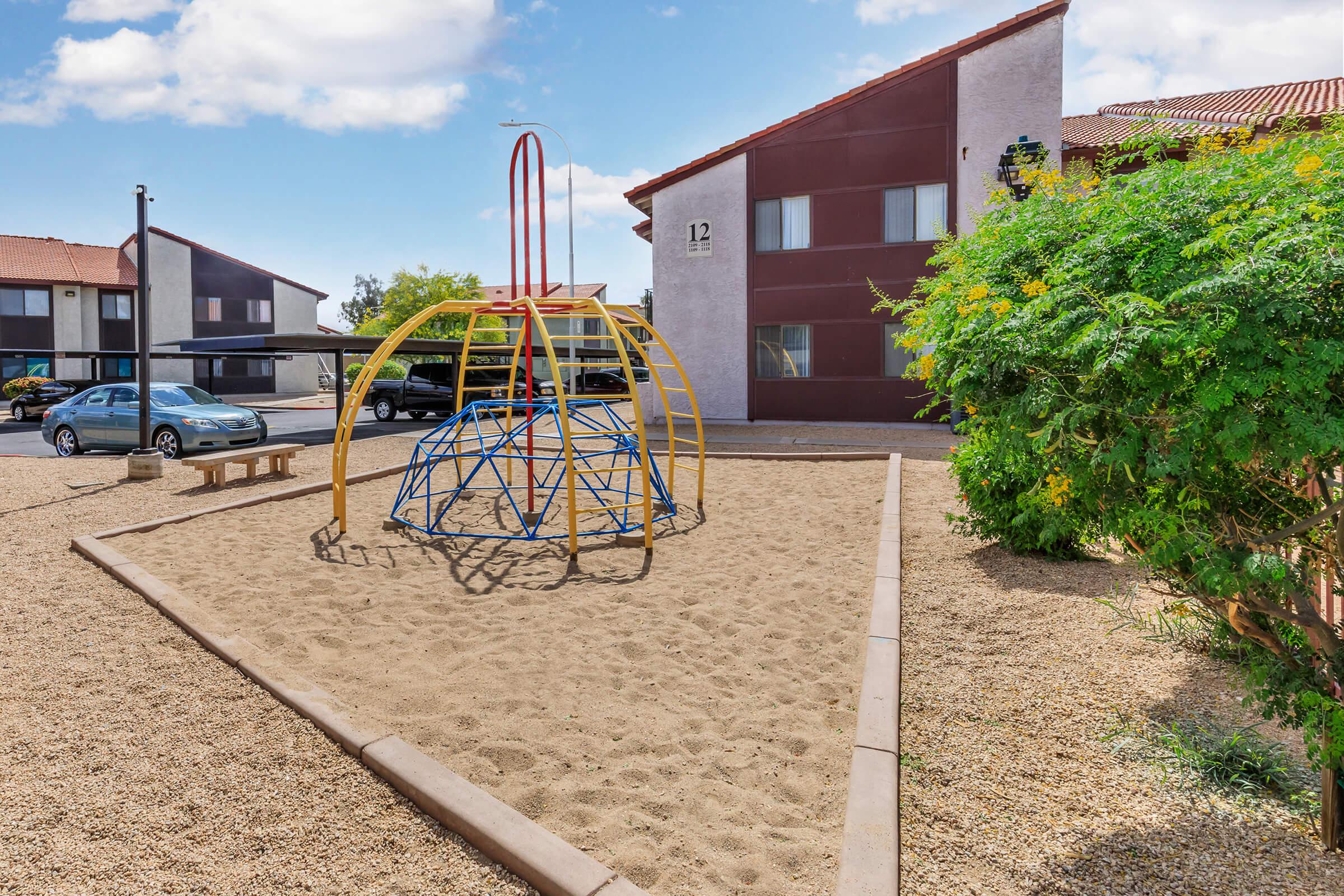 Playground area featuring a colorful climbing structure and sandy ground, surrounded by a landscaped garden. Nearby, there is a bench and residential buildings in the background. The scene is bright and sunny, creating a welcoming environment for children to play.