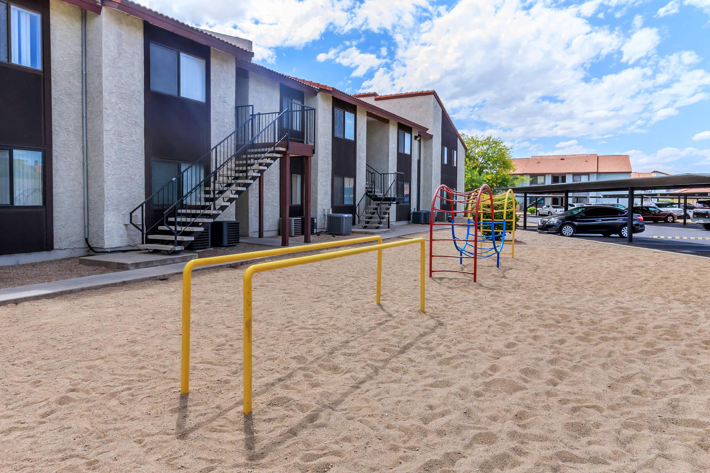 A playground area with yellow monkey bars and colorful climbing structures on a sandy surface, situated next to an apartment building. Staircases lead to the upper floors of the building, and there are parked cars nearby under a carport. The sky is partly cloudy.
