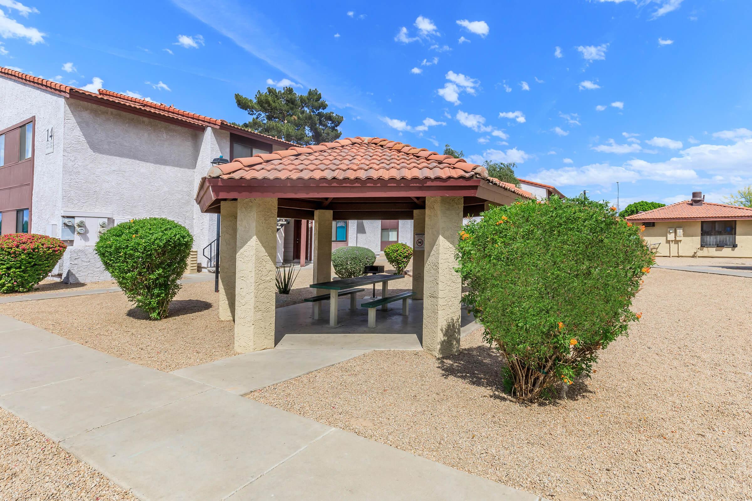 A shaded gazebo with a tile roof is set in a landscaped area, surrounded by low shrubs. The scene features a clear blue sky with a few clouds, and nearby buildings with a light-colored exterior. The ground is covered with small gravel, creating a neat outdoor space for relaxation.