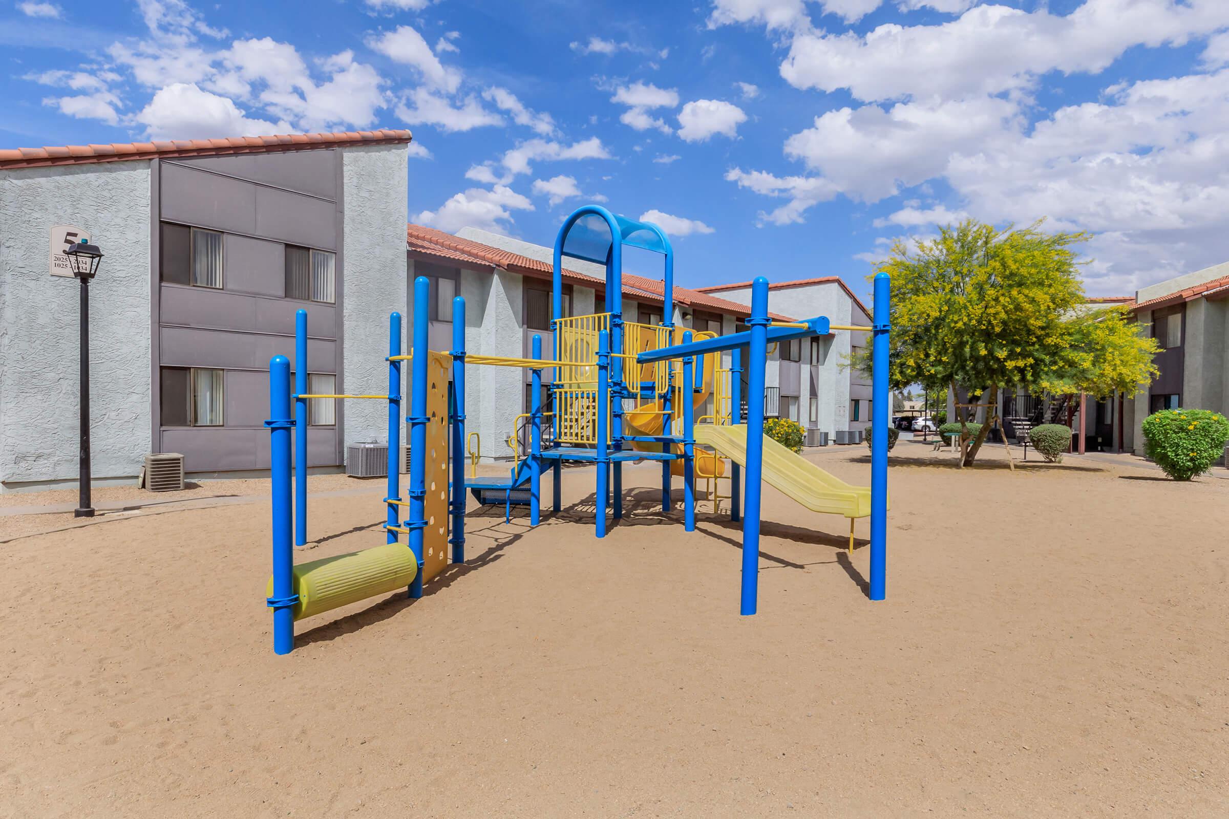 Colorful playground equipment featuring slides and climbing structures, set in a sandy area surrounded by low-rise apartment buildings and lush greenery under a blue sky with fluffy white clouds.