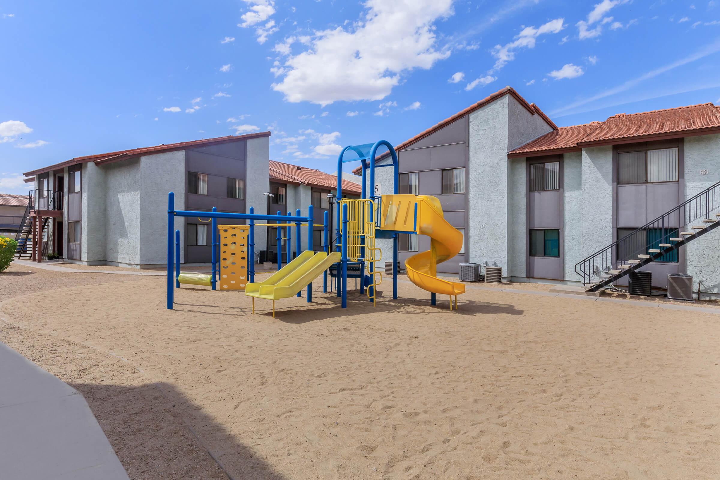 Playground with yellow and blue slides and climbing structures in a sandy area, surrounded by two-story apartment buildings with a clear blue sky and scattered clouds in the background.