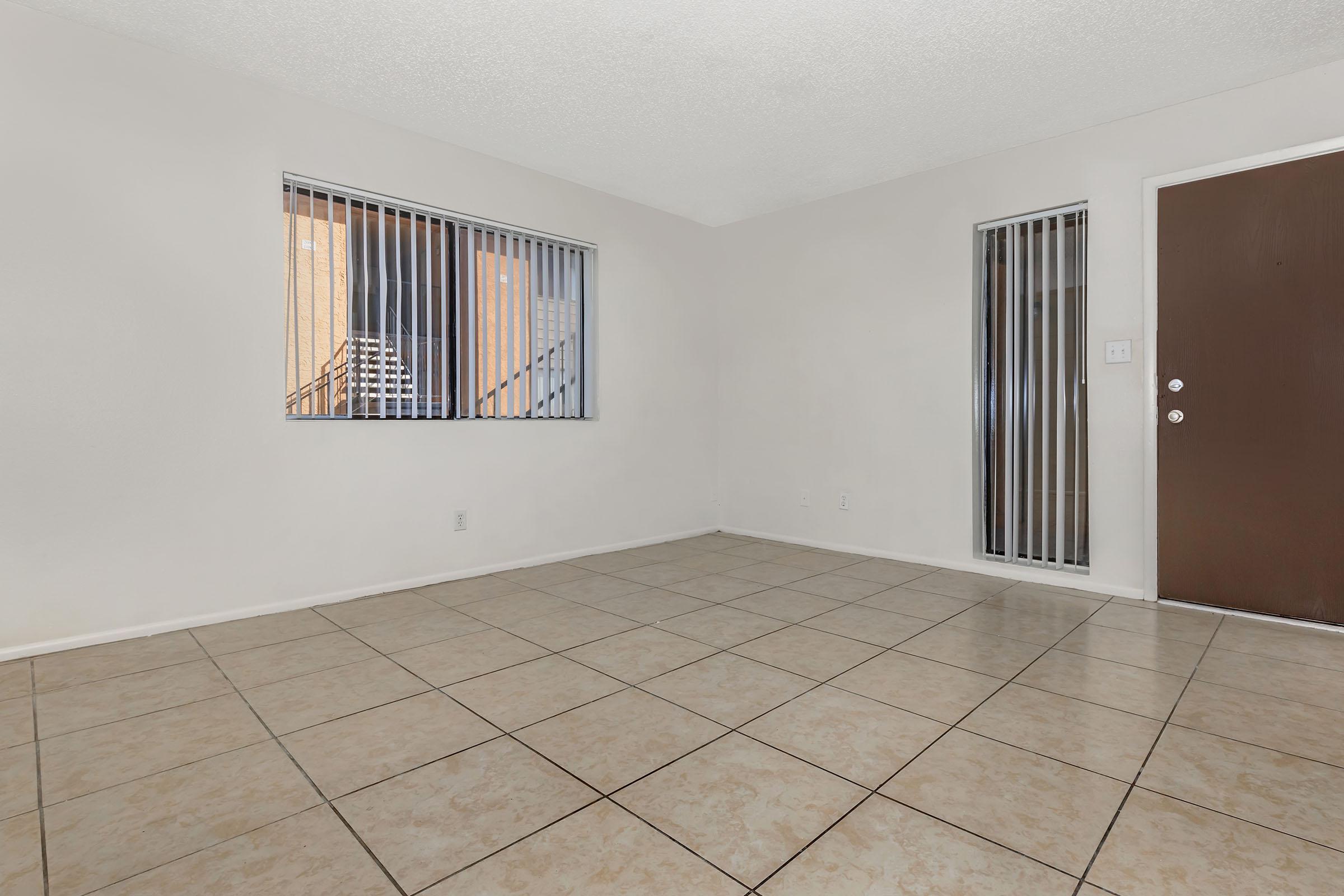 Empty room with ceramic tiled floor and light-colored walls. Features include vertical blinds on two windows, an exterior door with a modern handle, and no furniture or decorations present. Natural light enters through the windows, creating a bright and open atmosphere.