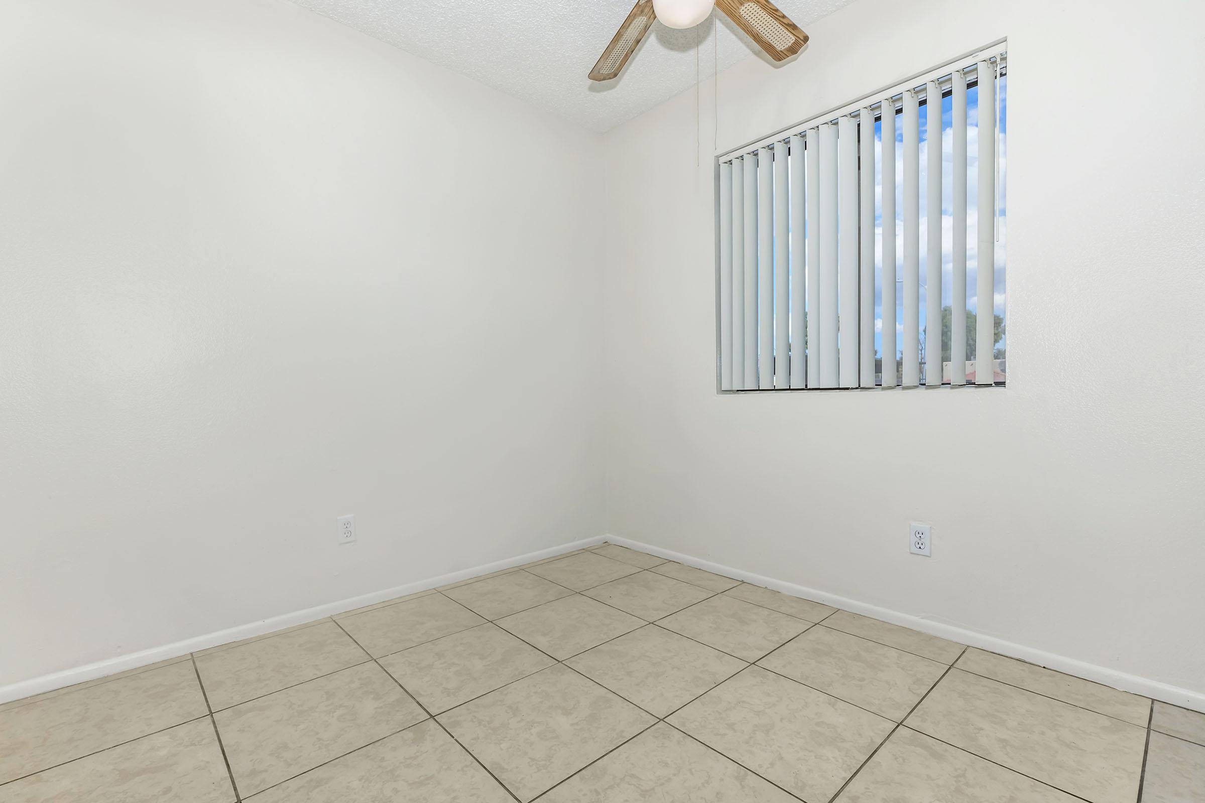 A empty room with light-colored walls and a tiled floor. It features a ceiling fan and a large window with vertical blinds, allowing natural light to enter. The corner of the room is visible, creating a spacious feel.