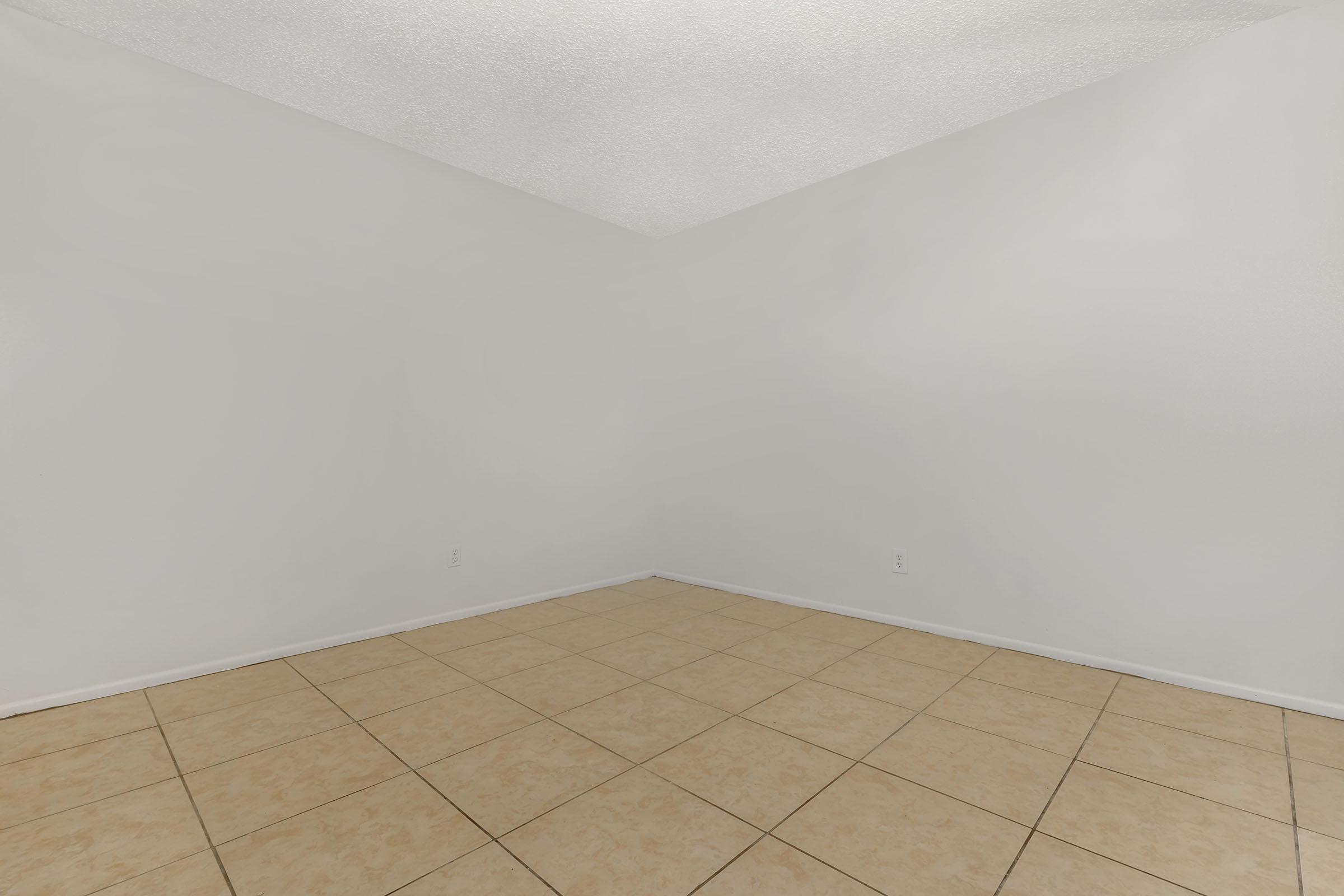 Empty room with light-colored walls and a tiled floor. One corner of the room is visible, showcasing the lack of furniture or decor. The ceiling has a textured finish, and the overall space appears bright and clean with natural light.