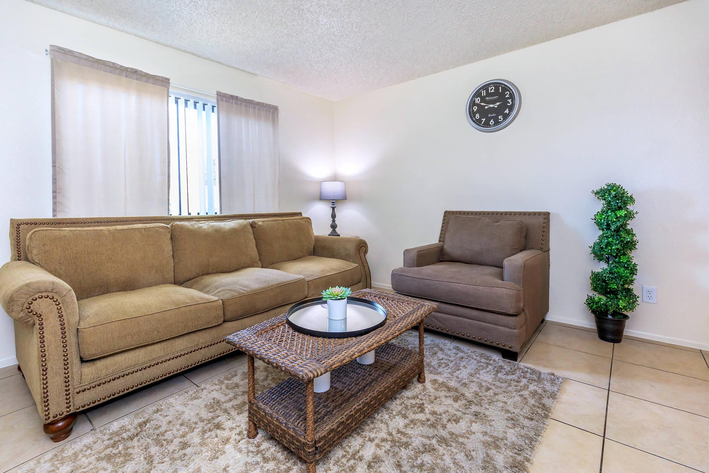 A cozy living room featuring a beige couch and an armchair, a round coffee table with a small plant, and a black wall clock. Natural light comes in through sheer curtains. The floor is tiled, and there is a decorative plant in the corner, creating a warm and inviting atmosphere.