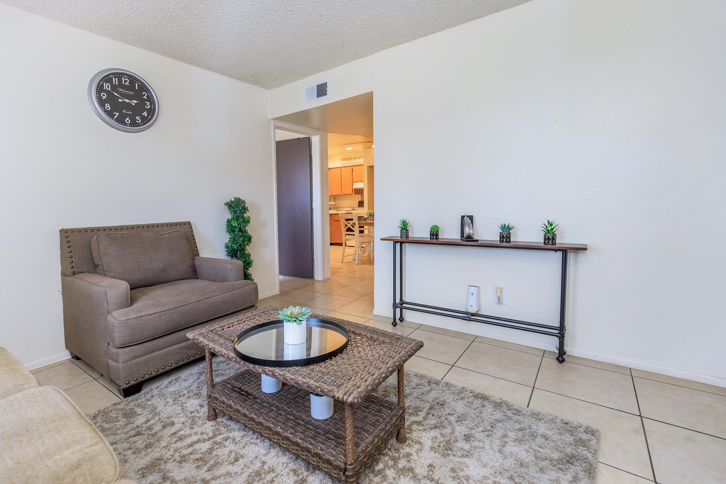Living room scene featuring a beige sofa and a gray armchair, a round coffee table with a plant centerpiece, and a decorative console table with small plants. A large wall clock is displayed above the armchair, and the space has light-colored tiled flooring. Natural light brightens the room from an open doorway.