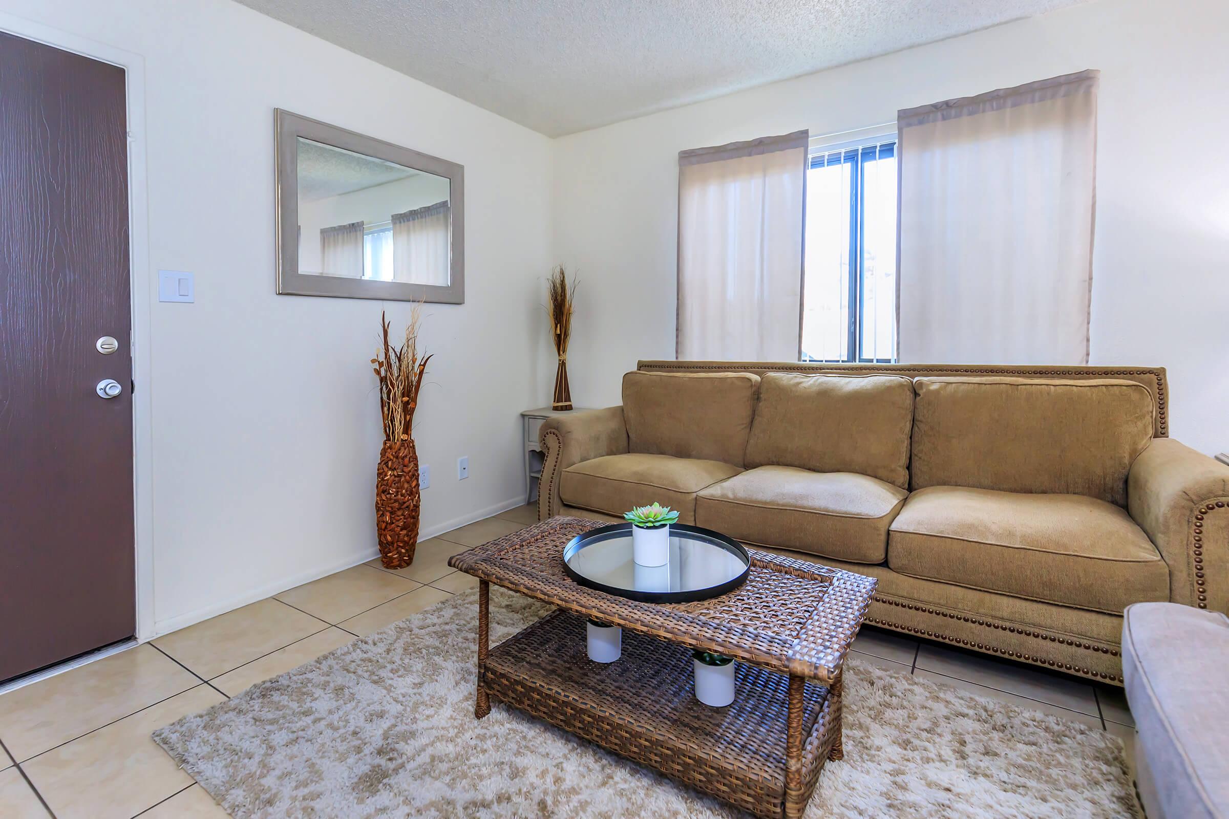 A cozy living room featuring a beige sofa, a glass-top coffee table on a woven mat, and decorative plants. Natural light filters through sheer curtains on two windows. A tall vase with dried plants adds a touch of elegance, while a large mirror reflects the space. The walls are painted in a light, neutral color.