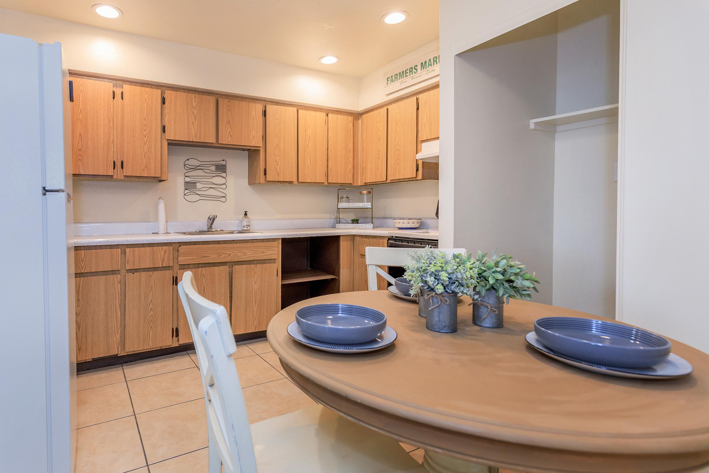 A cozy kitchen featuring wooden cabinets, white countertops, and a small dining table set for two with gray plates. There is a refrigerator, a stove, and a sink visible, along with a decorative plant on the table and an open shelf in the corner. The space is well-lit with overhead lights.