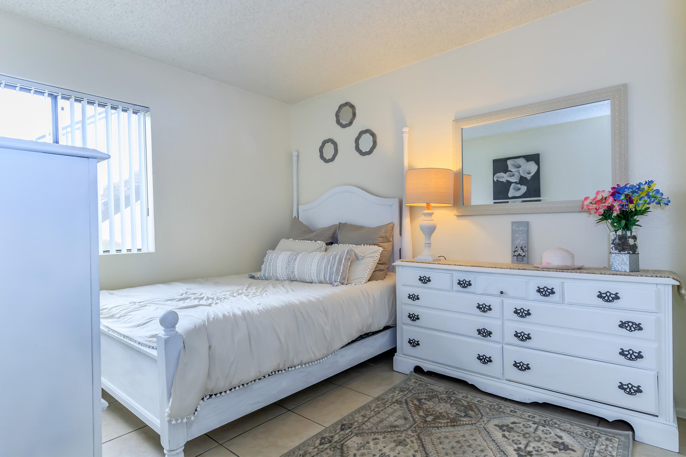 A cozy bedroom featuring a light-colored bed with decorative pillows, a white dresser with ornate handles, and a mirror on the wall. There's a lamp providing warm light, and a vase of colorful flowers sits on the dresser. The room has a serene atmosphere with neutral tones and a tiled floor.