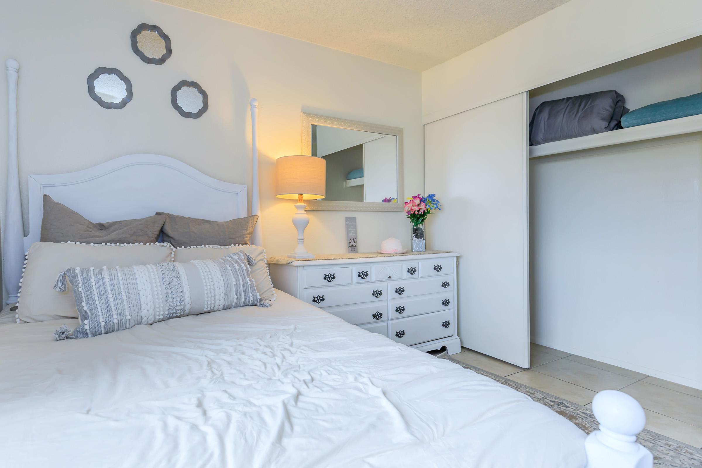 A cozy bedroom featuring a white bed with decorative pillows, a bedside table with a lamp, a mirror above the dresser, and a closet with neatly arranged items. There are three round mirrors on the wall, and a bouquet of flowers adds a touch of color to the room.
