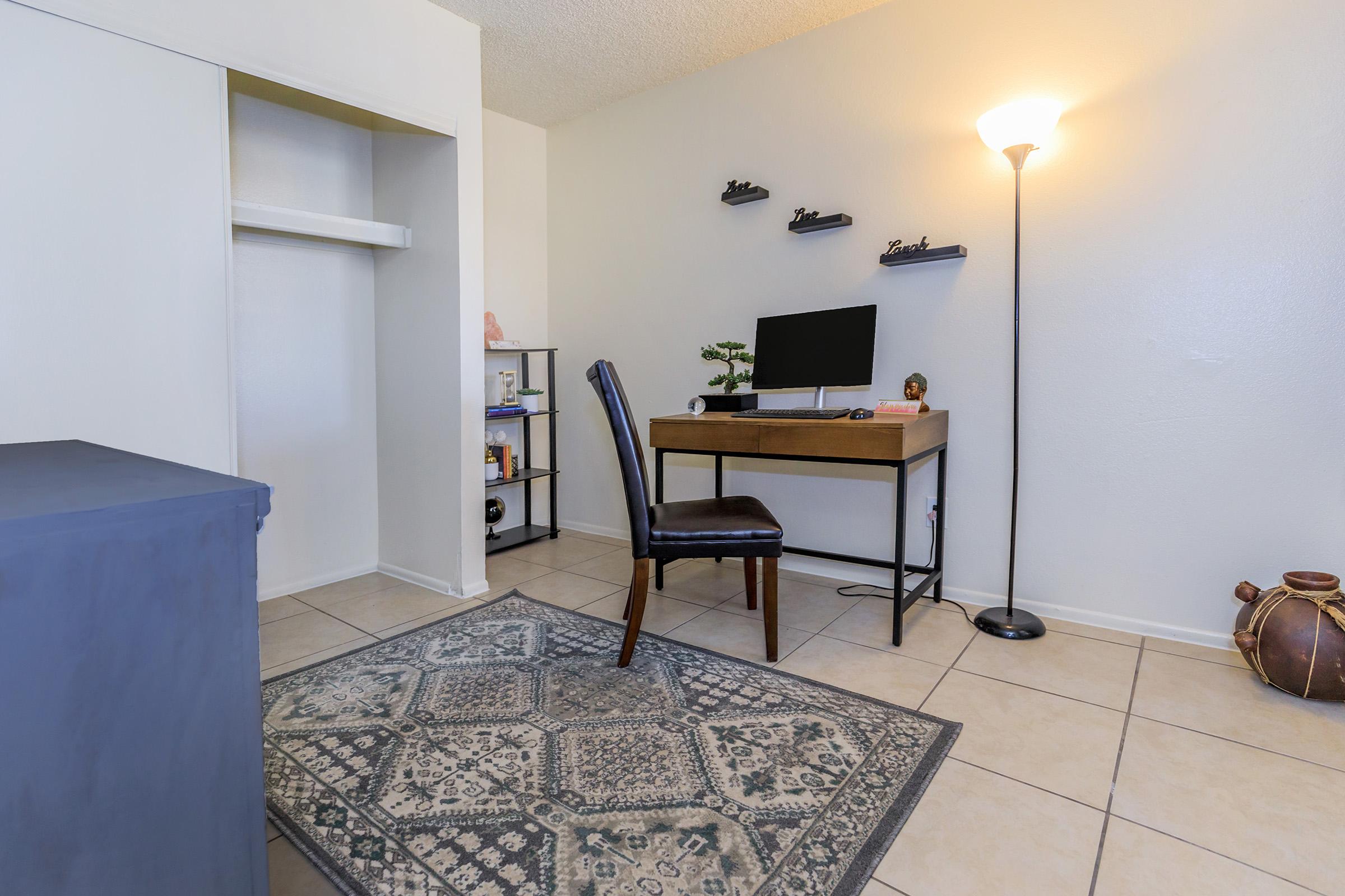 A small, minimalist home office featuring a wooden desk with a computer monitor, a black chair, and decorative items on shelves. A tall floor lamp provides light, and a patterned area rug adds comfort. A closet is partially visible on the left, and a decorative pot is placed in the corner.