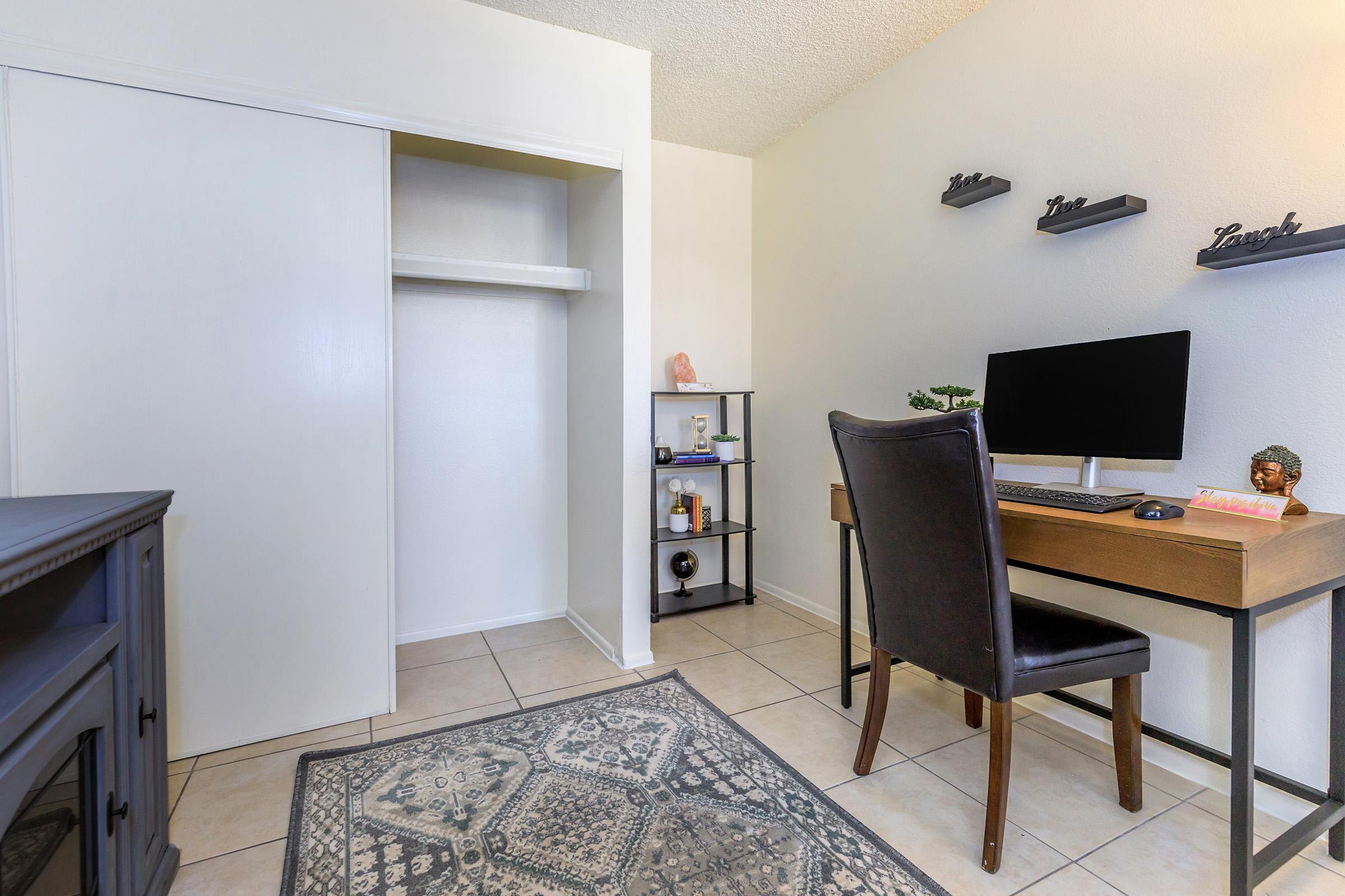 A small office space featuring a desk with a computer and a comfortable black chair. There is a shelving unit with decorative items and a closet with sliding doors on one side. The room has light-colored walls and tiled flooring, creating a clean and minimalistic atmosphere.