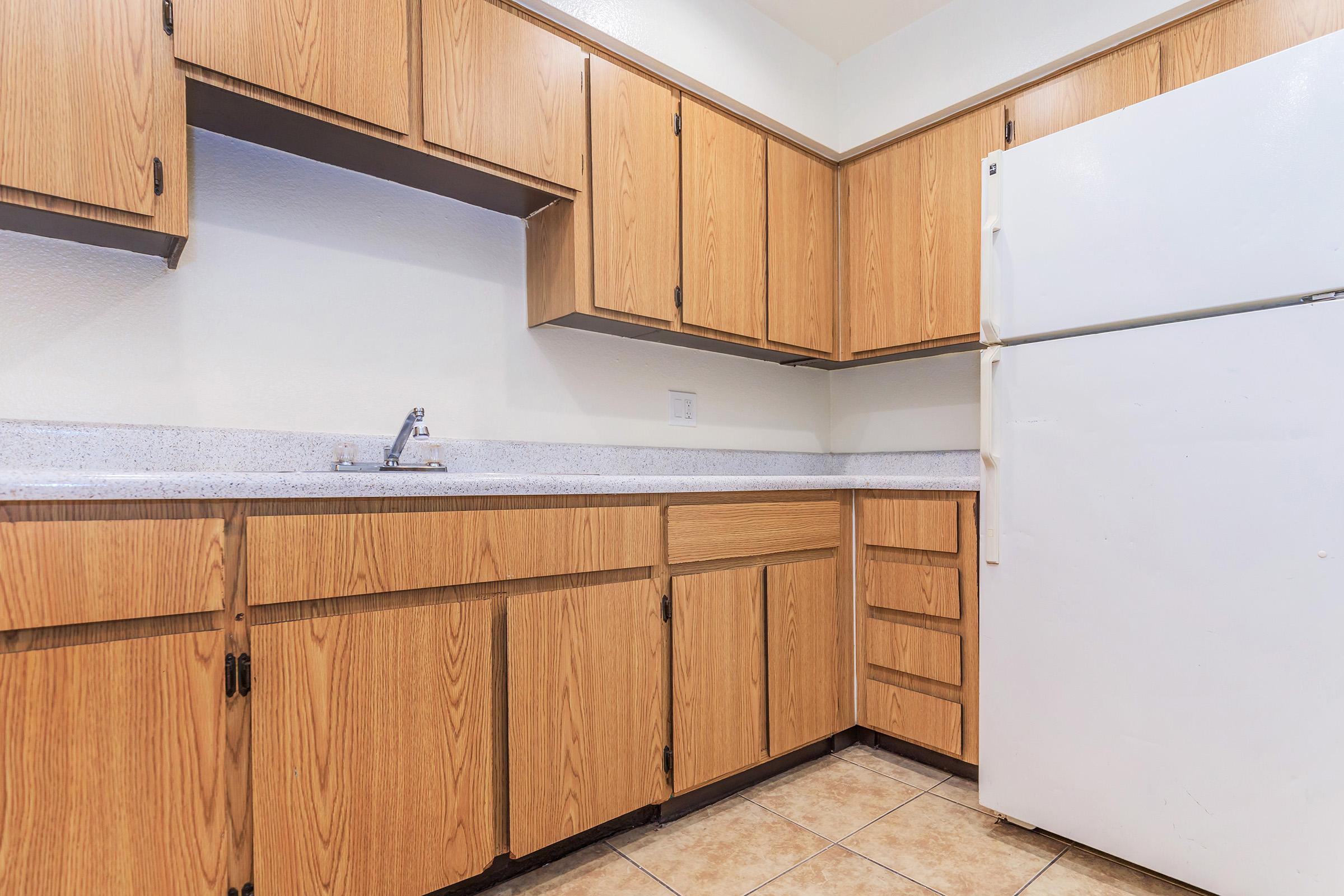 A kitchen with light brown wooden cabinets, a countertop, and a white refrigerator. The space features a simple design with a sink and several drawers, all set against a neutral-colored wall and tiled floor. The overall atmosphere is clean and functional.