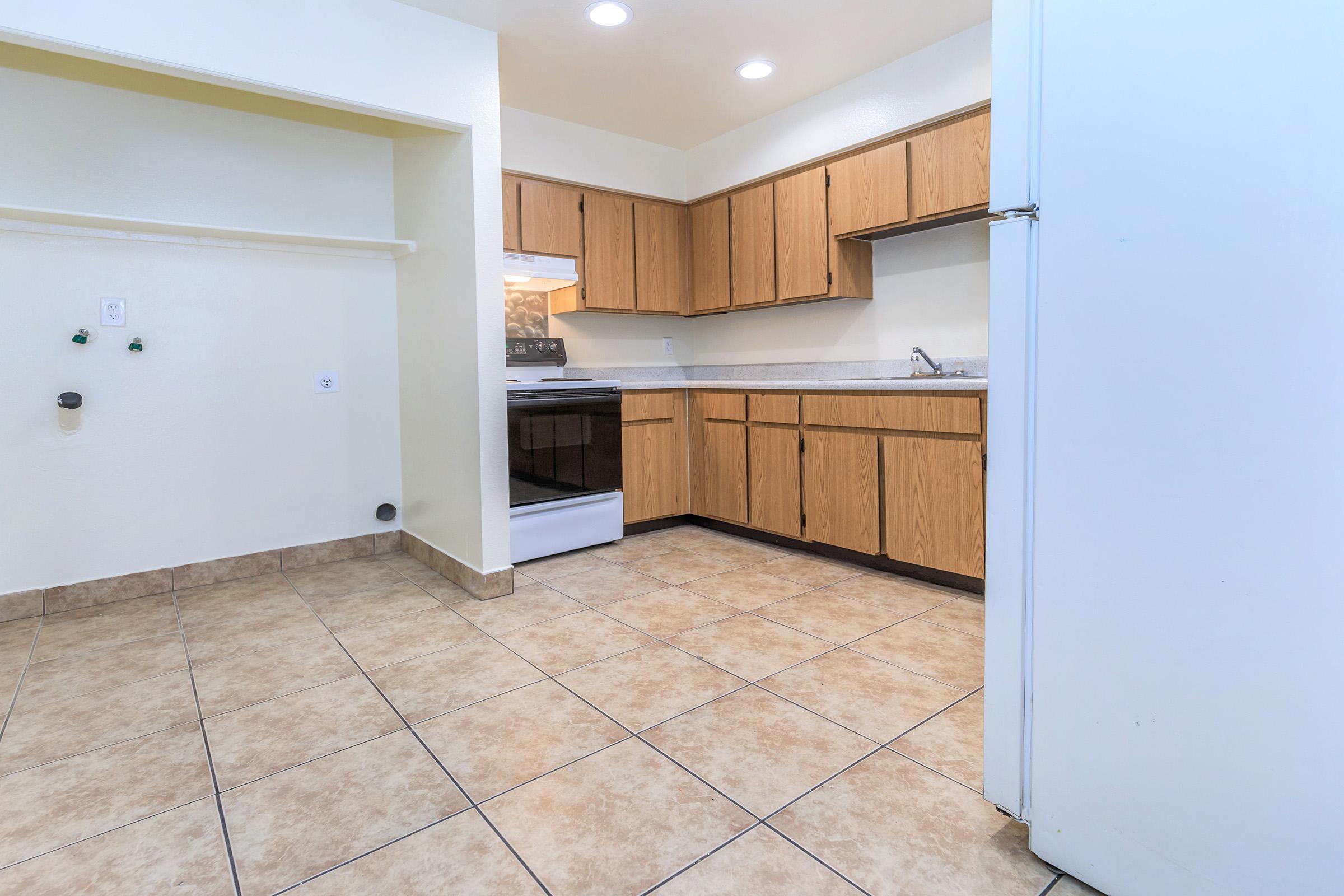 Bright, empty kitchen featuring tan tiled flooring, wooden cabinets, and a white stove. The layout includes a sink and countertop, with natural light coming from a nearby window. The space is clean and uncluttered, making it ideal for cooking and dining.