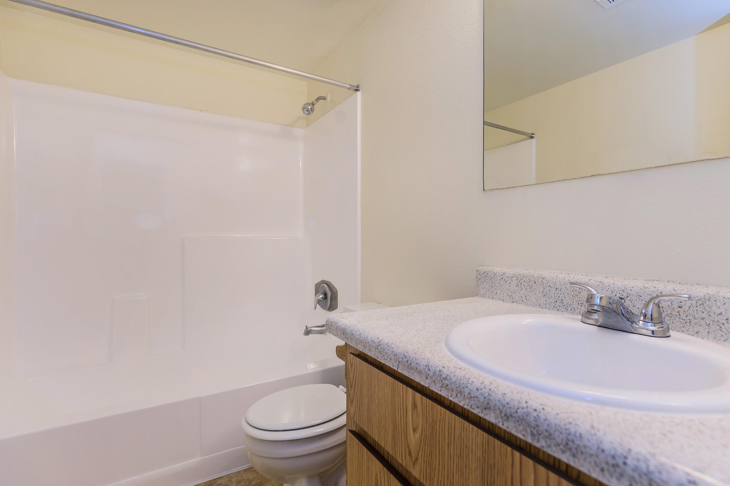 A clean, minimalist bathroom featuring a white shower/tub combo, a light-colored countertop with a sink, and wooden cabinetry. The wall is painted in a light shade, and there is a large mirror above the sink reflecting the space. The flooring is beige tile.