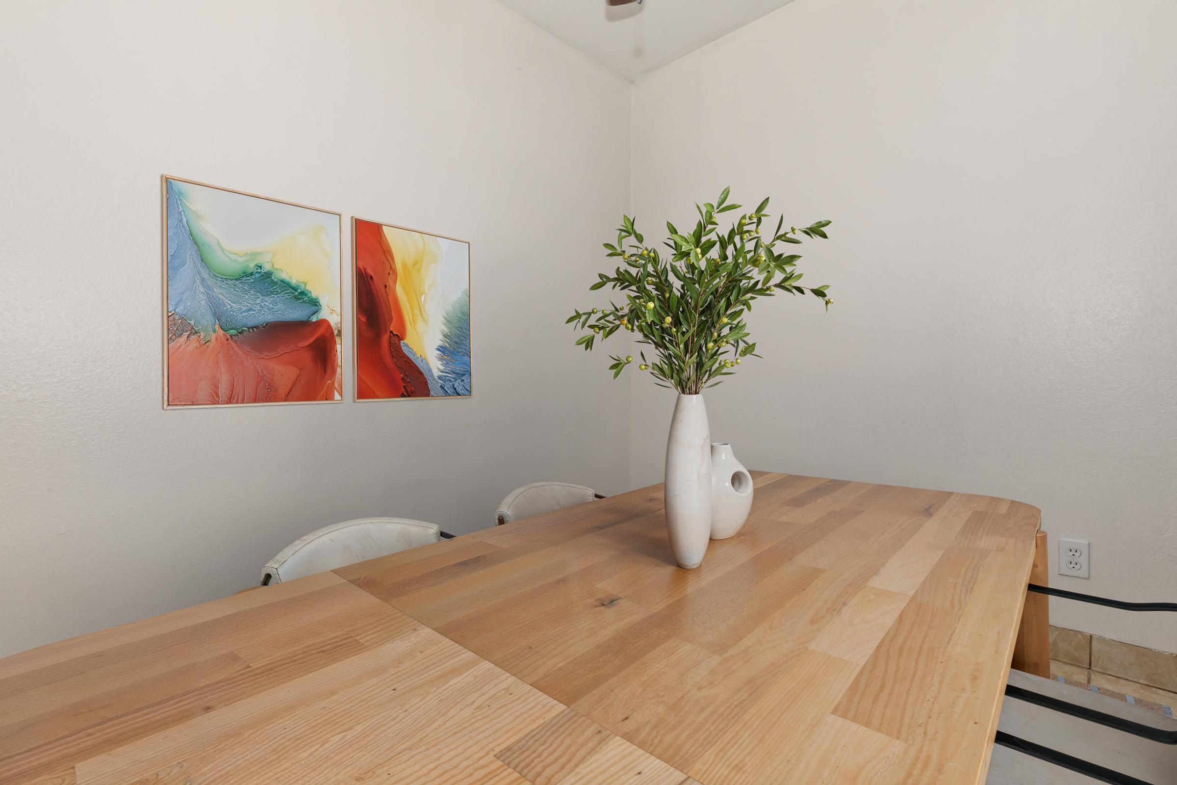 A modern dining area featuring a wooden table with a vase of greenery on it. The wall is adorned with two colorful abstract paintings in blue, red, yellow, and green. The space has a minimalist design and a warm, inviting atmosphere.