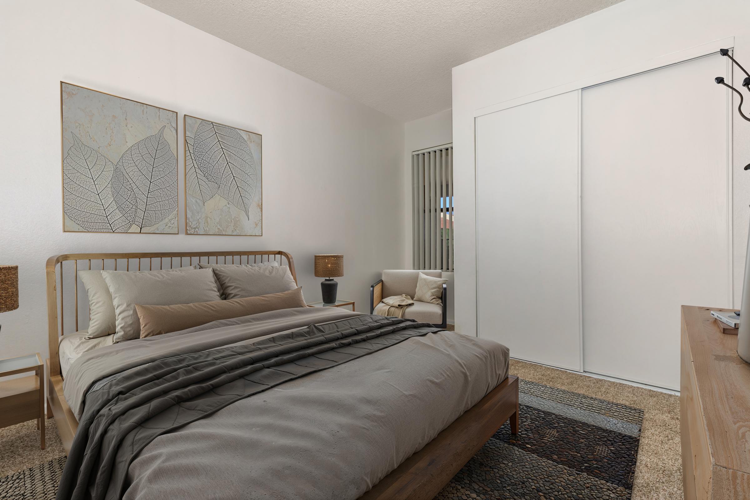 A cozy bedroom featuring a wooden bed with neutral-colored bedding, two leaf-patterned wall art pieces, and bedside tables with lamps. A comfortable chair is positioned near a sliding closet door, and textured rugs add warmth to the space. Natural light filters through a window with blinds.