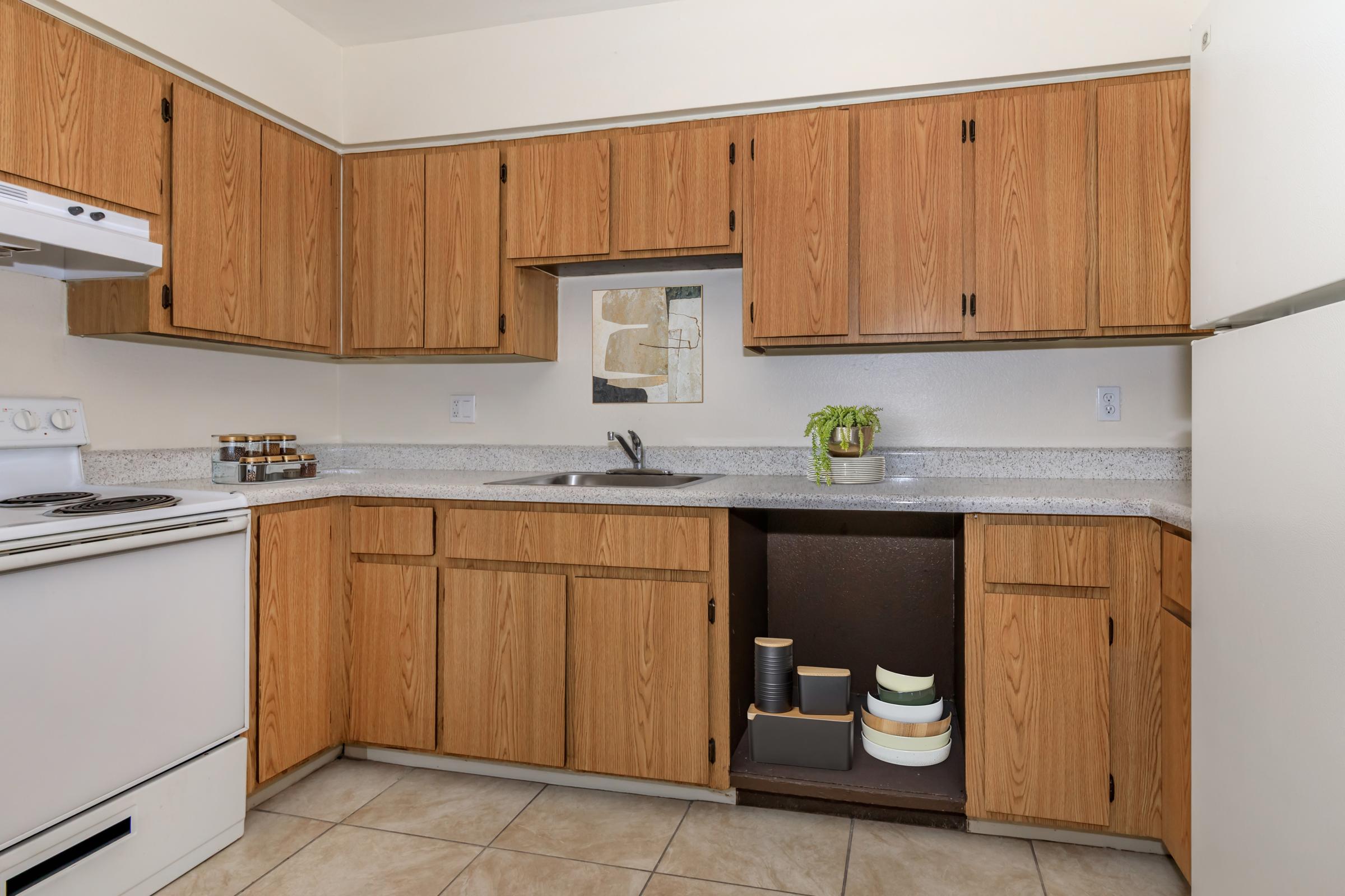A modern kitchen featuring wooden cabinets, a white stove and refrigerator, a sink, and countertop space. There are jars on the counter, and a plant on the sink area. The floor is tiled, and the overall atmosphere is bright and functional.