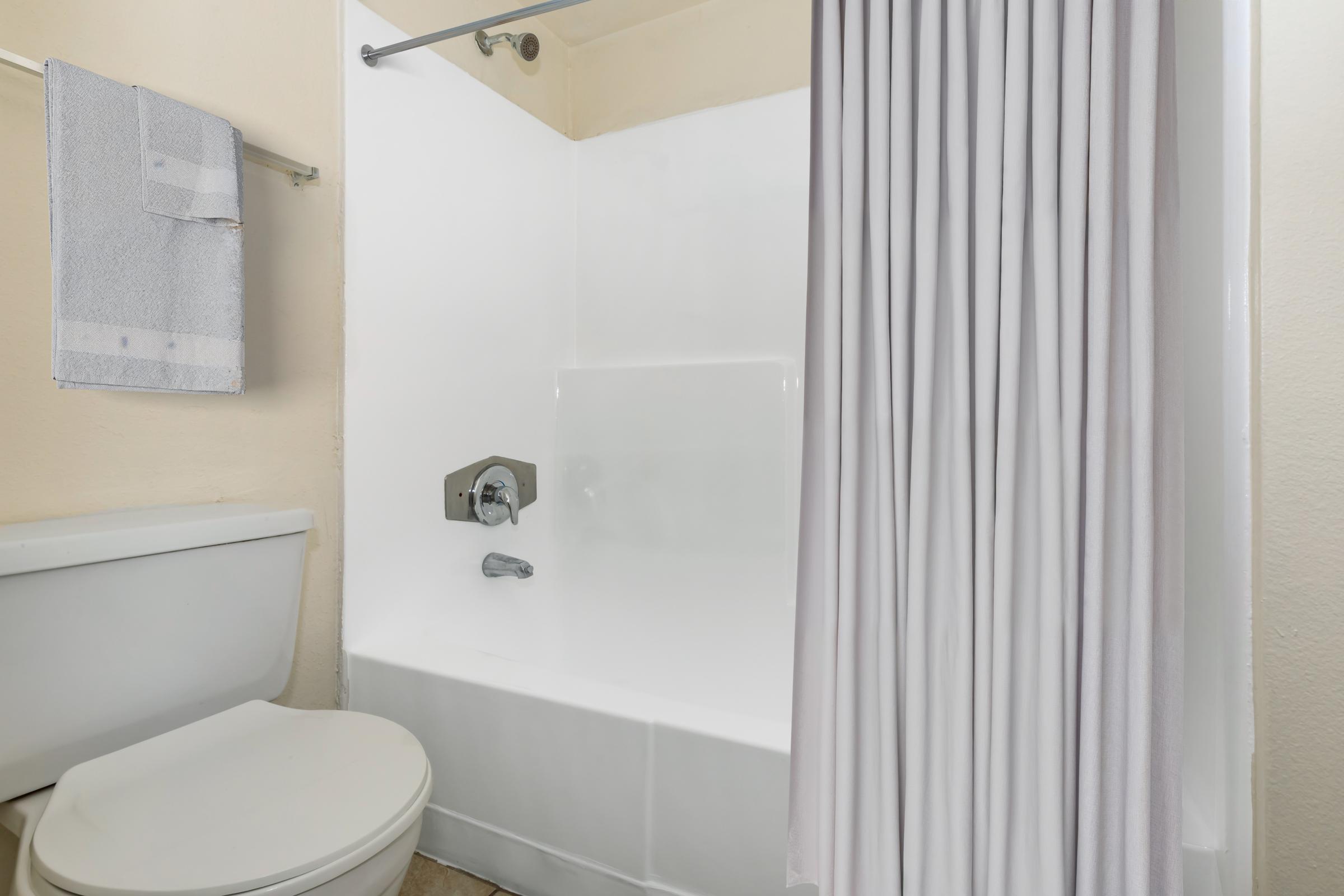 A simple bathroom with a white bathtub and shower combination. A white shower curtain is partially drawn, and a gray towel hangs on a towel bar nearby. The toilet is visible to the left of the bathtub, and the walls are painted a light color. The space appears clean and well-maintained.
