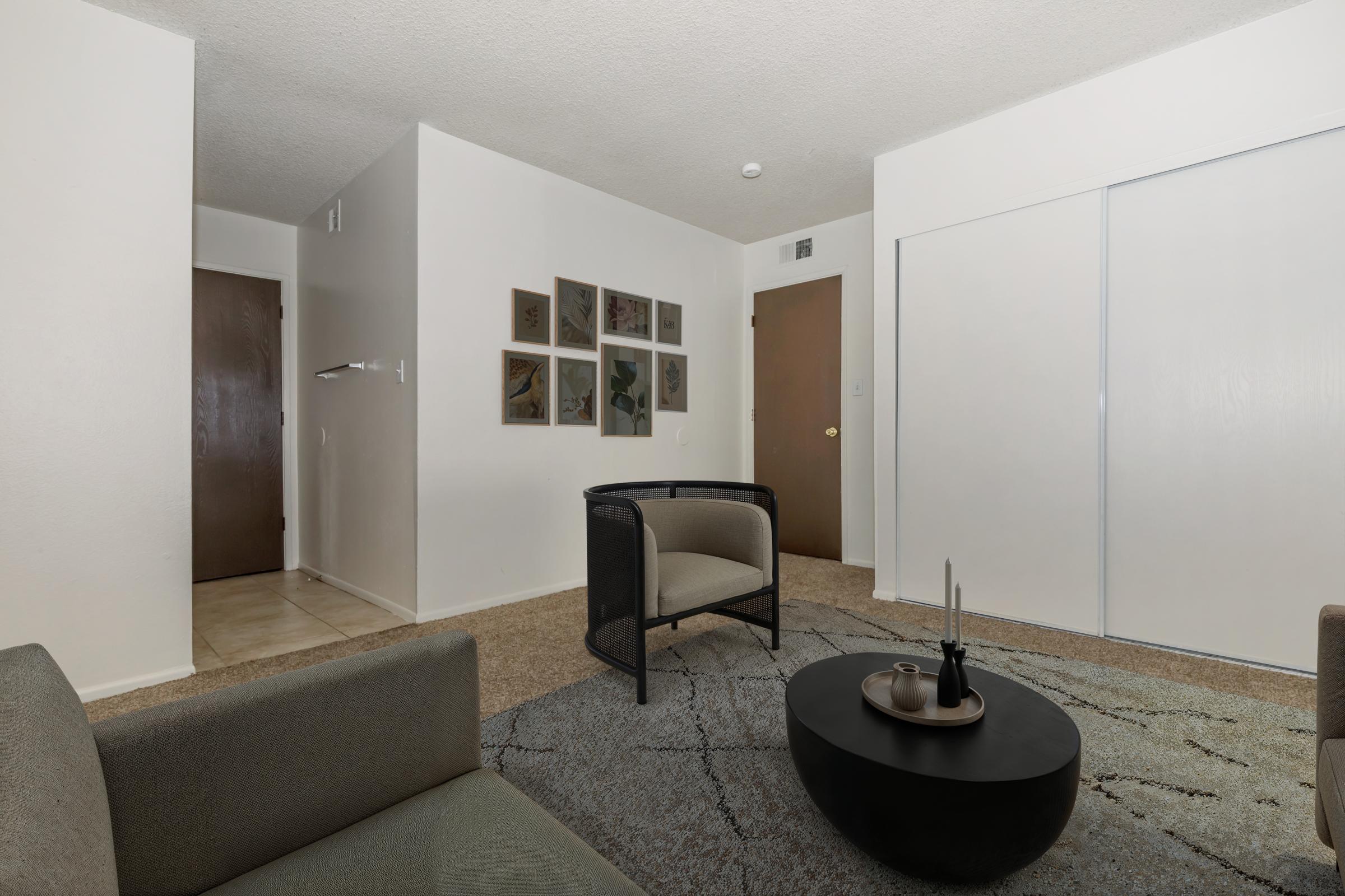 A modern living space featuring two armchairs and a round coffee table. The room has light-colored walls, tile flooring, and a textured area rug. A series of framed artworks is displayed on one wall, and a hallway with a wooden door is visible in the background. The atmosphere is cozy and inviting.
